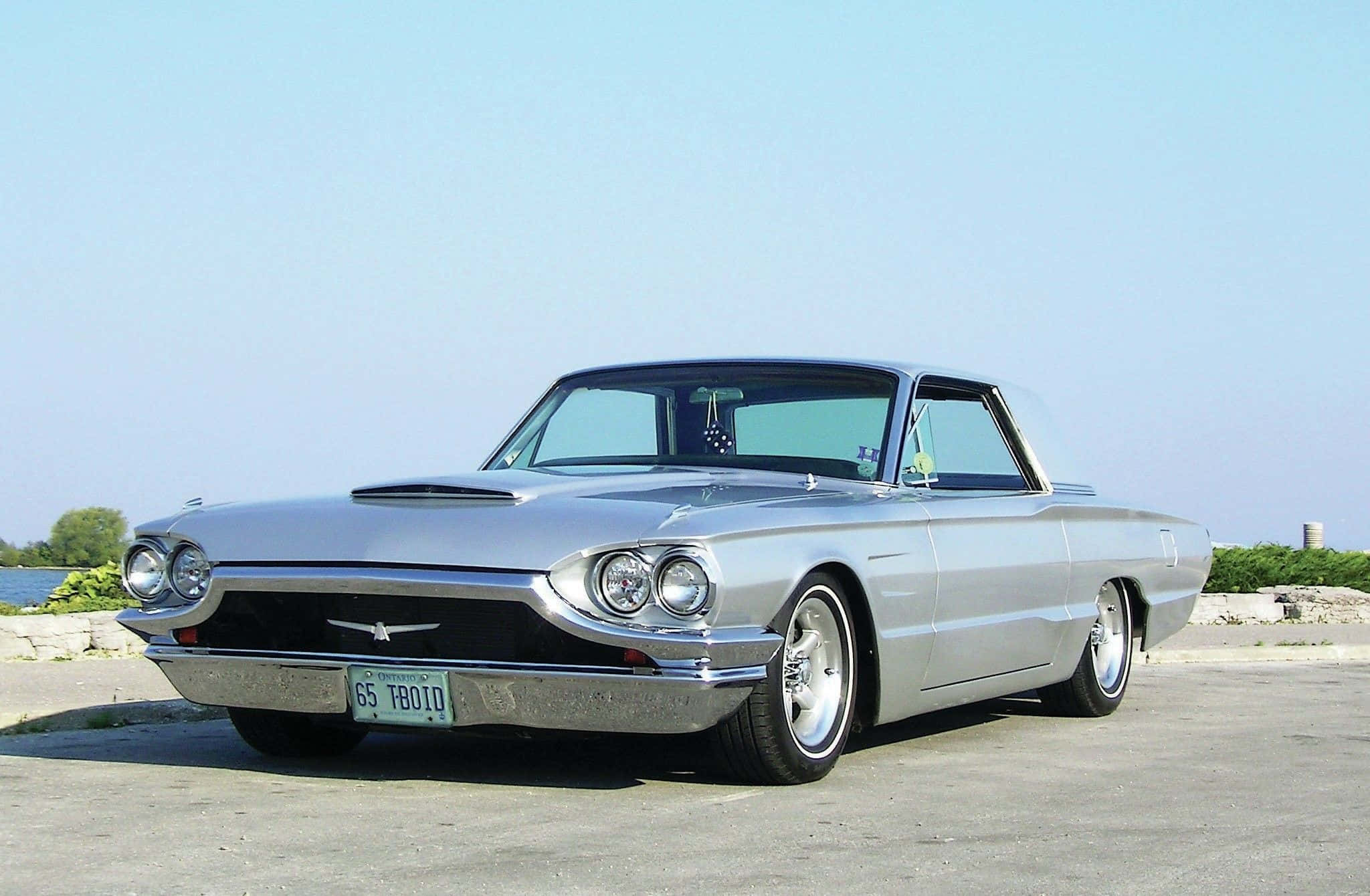 Ford Thunderbird 桌布 壁紙 — 一輛閃亮的銀色1965年福特雷鳥經典汽車停在瀝青路上，背景是平靜的湖泊和綠植，上方是清澈的藍天。