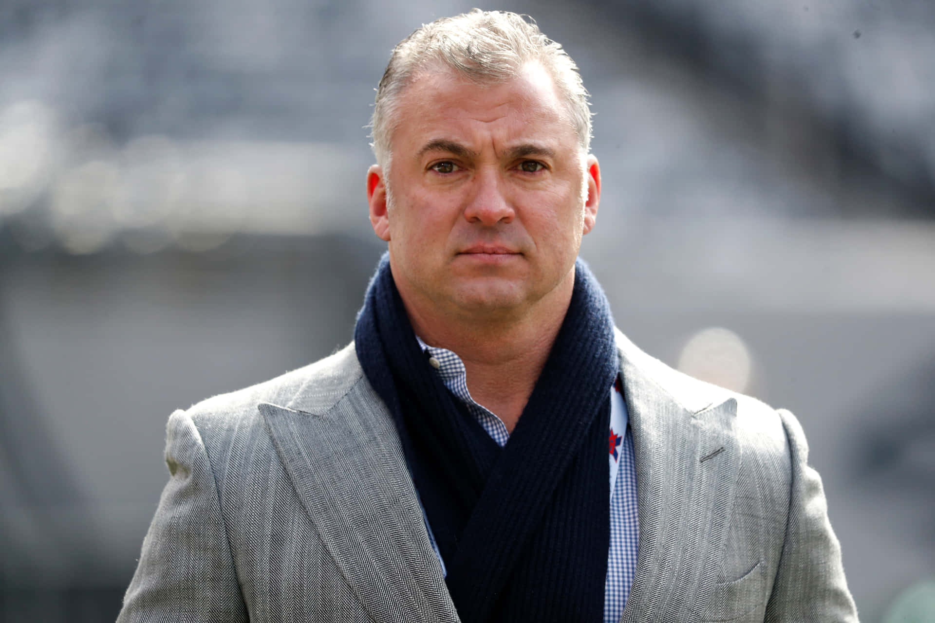 Shane McMahon 在行動中 — 美國商人的藝術 壁紙 — 一名身穿灰色西裝和深藍色圍巾的男子，在模糊的體育場背景下表情嚴肅地站立。