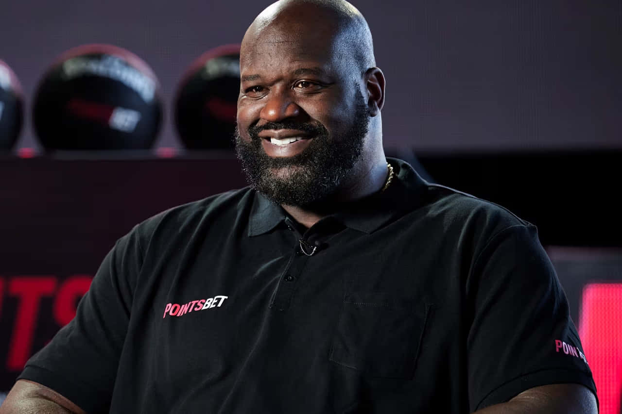  壁紙 — Shaquille O'Neal 穿著黑色Polo衫，上面有'POINTS BET'標誌，在深色背景中與籃球一起溫暖微笑。