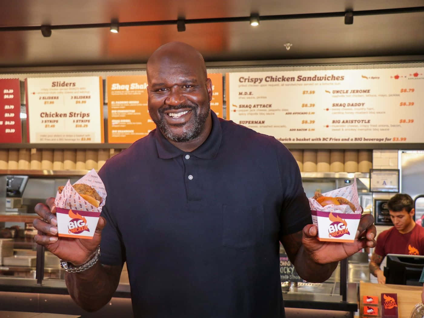  壁紙 — Shaquille O'Neal 喺快餐店嘅餐牌前，拎住兩個 Big Chicken 三文治，笑容滿面。