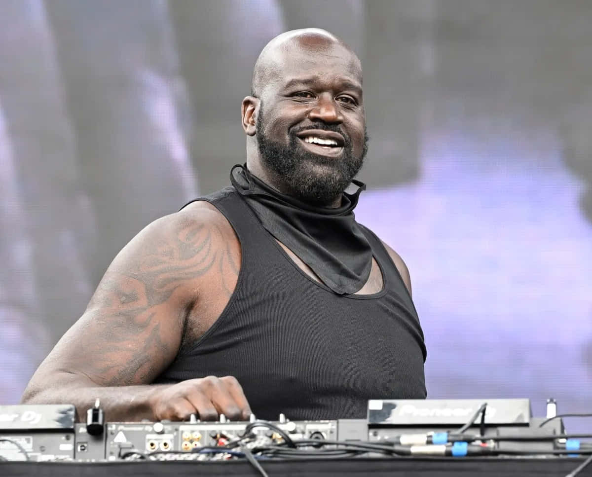  壁紙 — Shaquille O'Neal 喺戶外音樂會做DJ，露出紋身手臂，身穿黑色背心，咧嘴大笑。