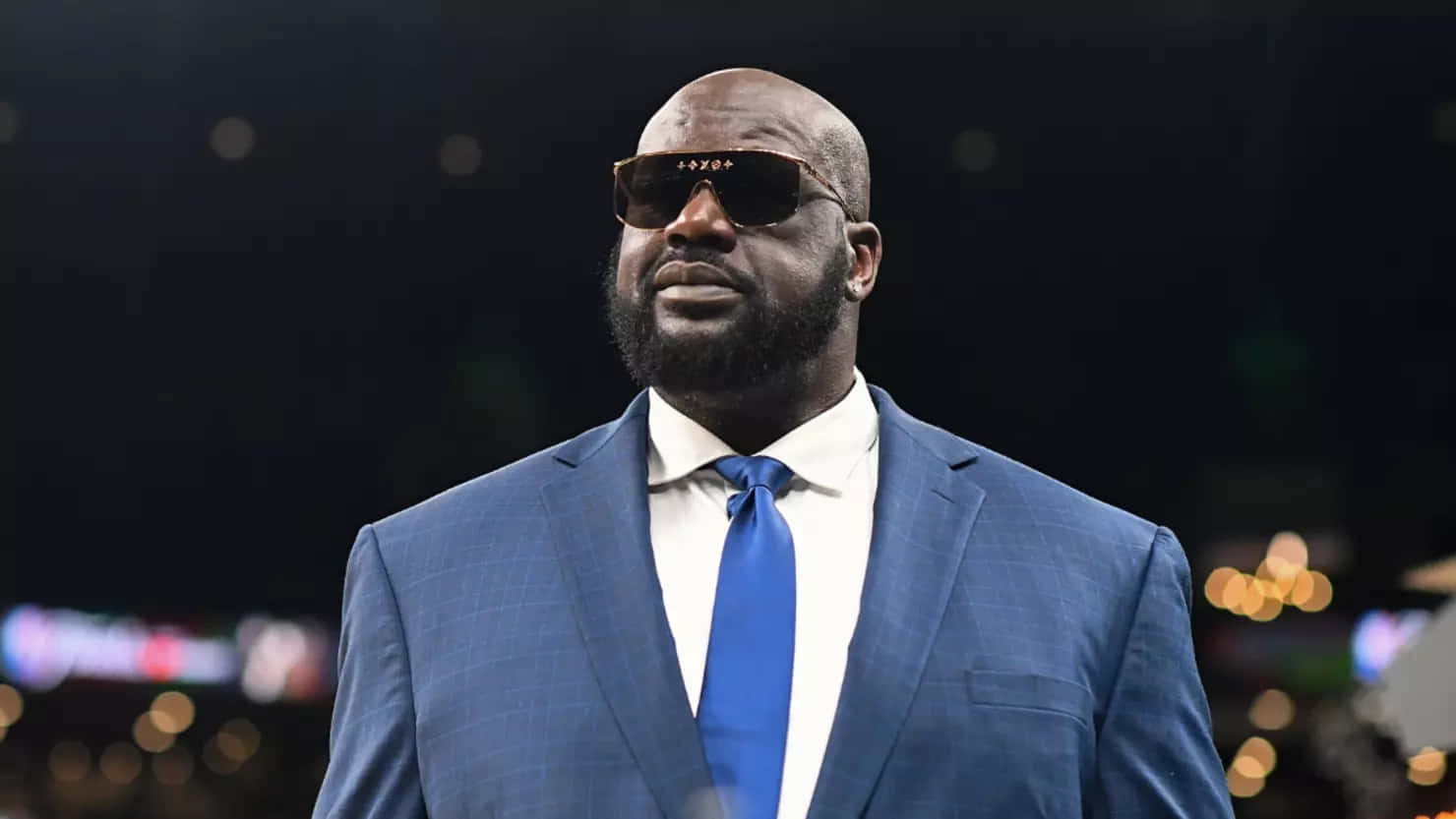  壁紙 — Shaquille O'Neal 身穿俐落藍色西裝和太陽眼鏡，自信地站在模糊的競技場背景前。