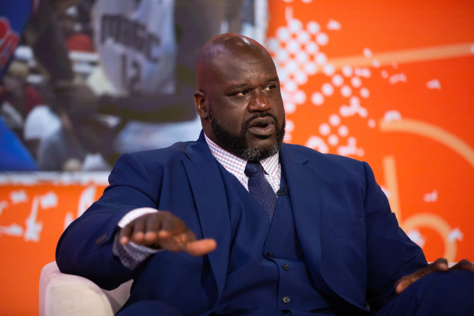  壁紙 — Shaquille O'Neal 身穿俐落藍色西裝，坐在白色椅子上，在帶有抽象白色圖案的鮮豔橙色背景前做手勢。