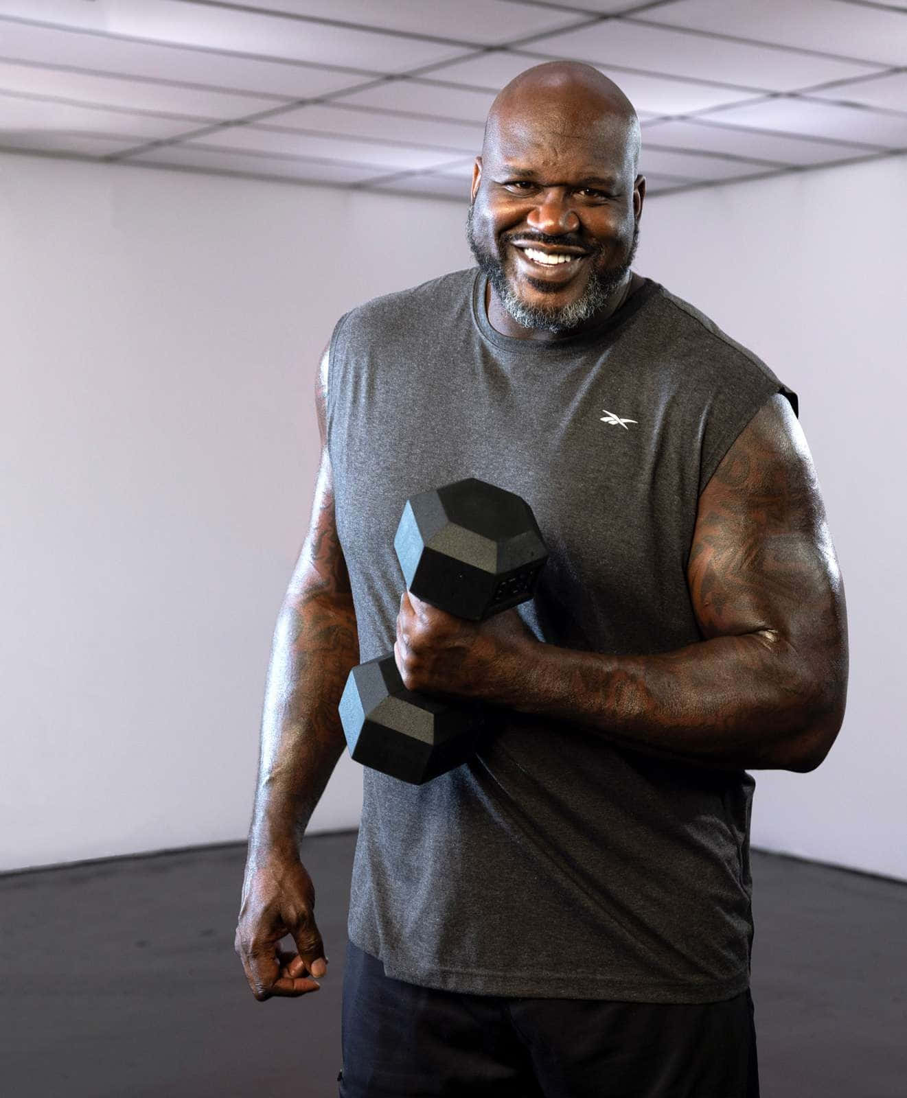  壁紙 — Shaquille O'Neal 在簡約的健身房環境中，手持黑色六角啞鈴，露出燦爛的笑容。