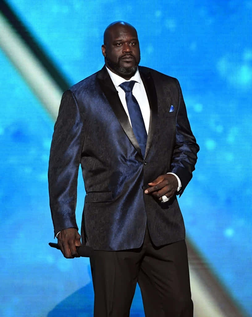  壁紙 — Shaquille O'Neal 自信地站在舞台上，身穿質感藍色天鵝絨禮服，背景是發光的藍色。
