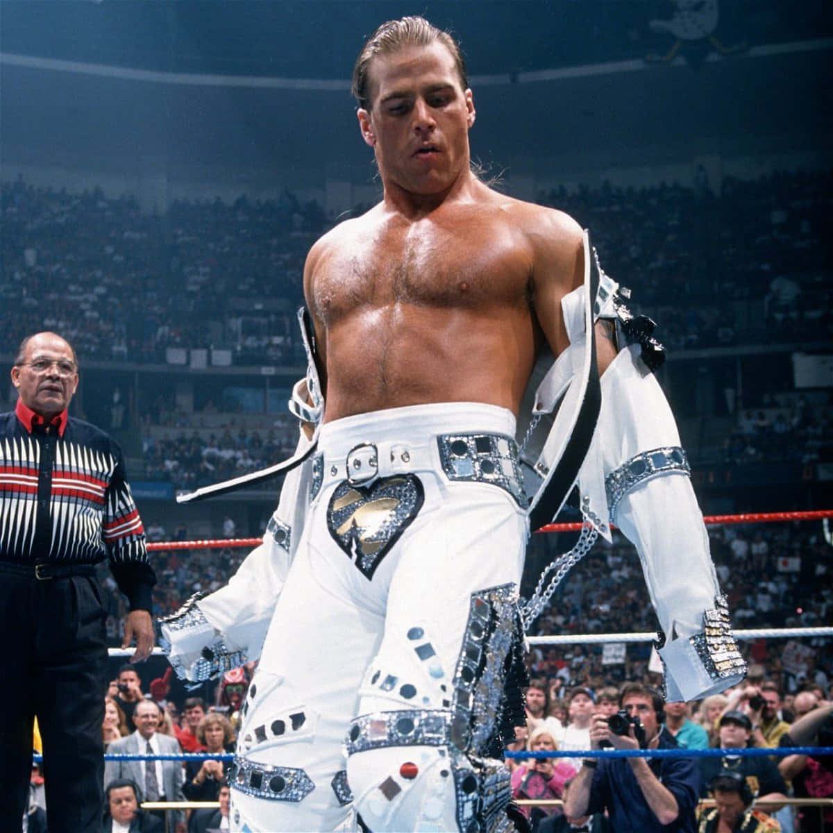 美國摔角手 Shawn Michaels 壁紙 — 一位肌肉發達的摔角手身穿白色閃片服裝，站在擂台上，裁判在他旁邊，觀眾在觀看。