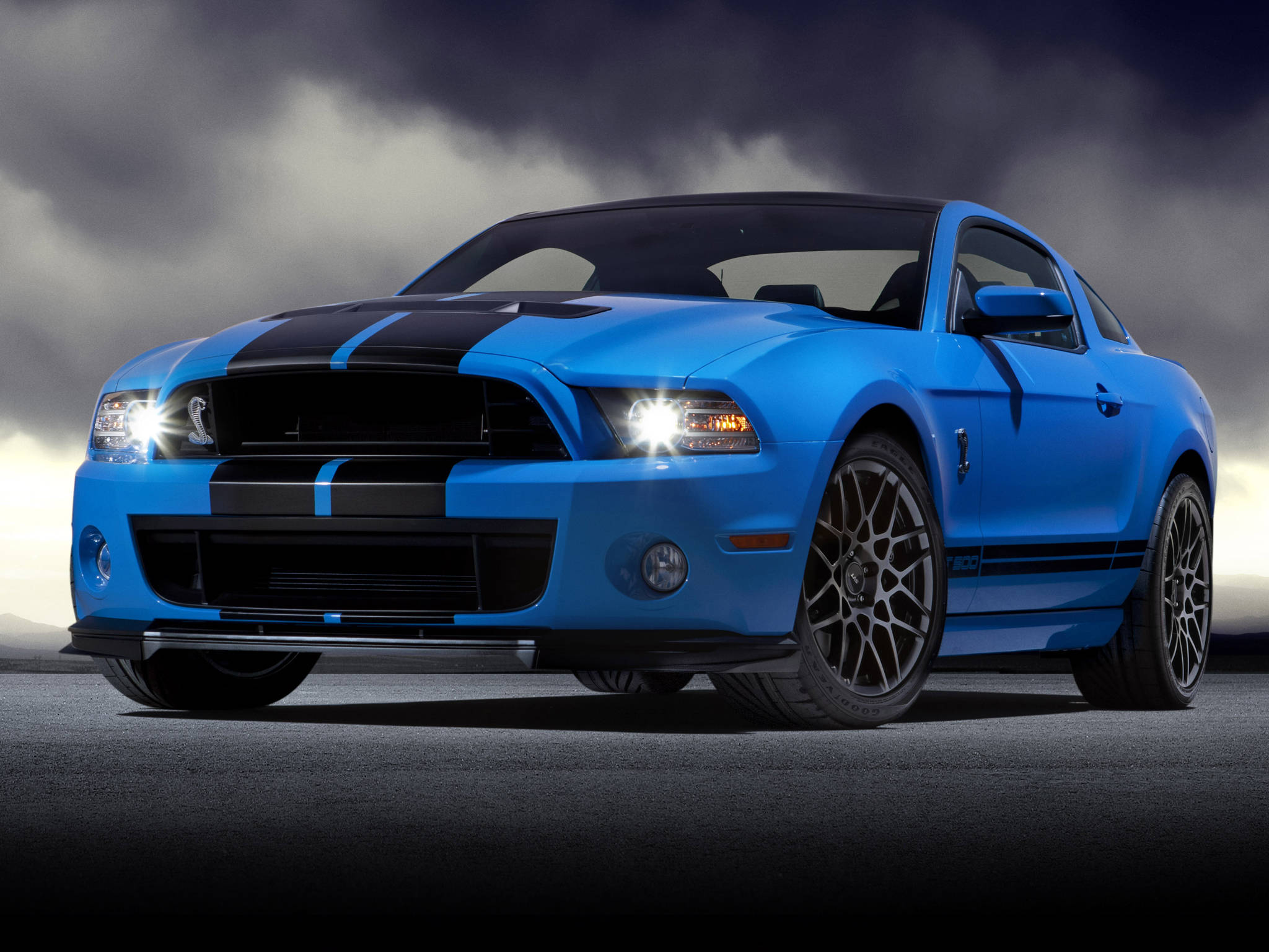 Shelby iPhone Ford Mustang 壁紙 — 一輛充滿活力的藍色福特野馬 Shelby GT500，帶有黑色賽車條紋，在戲劇性的暴風雲下亮著頭燈蓄勢待發。