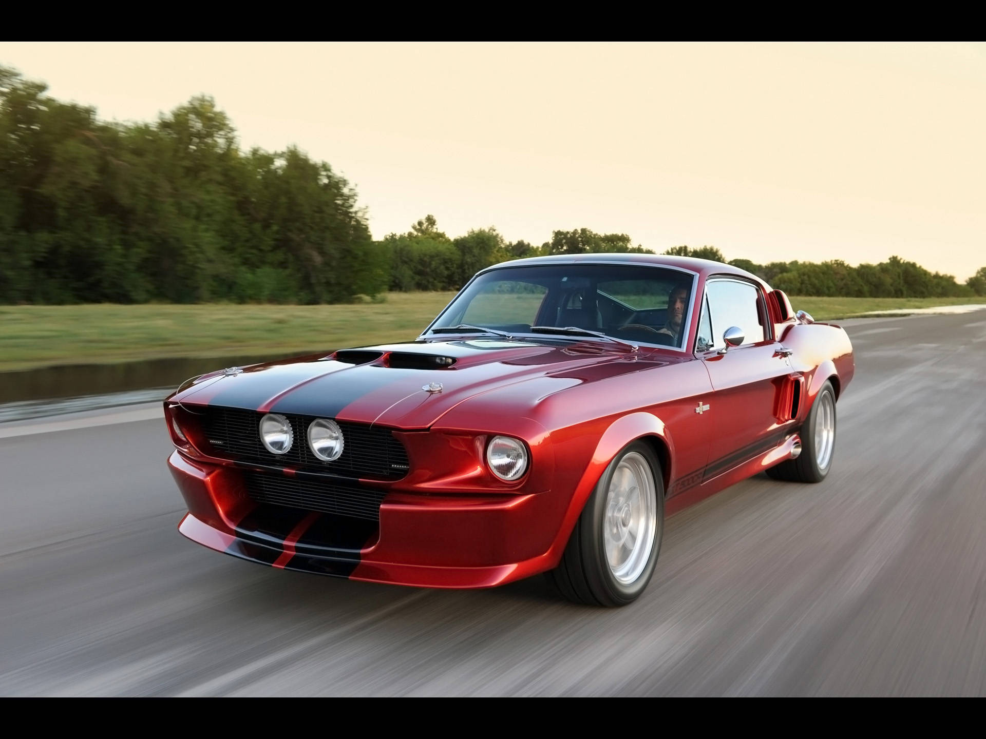 Shelby iPhone 金屬紅色款 壁紙 — 一輛鮮紅色的經典肌肉車帶有黑色賽車條紋，在日落時分沿著鄉村道路飛馳。