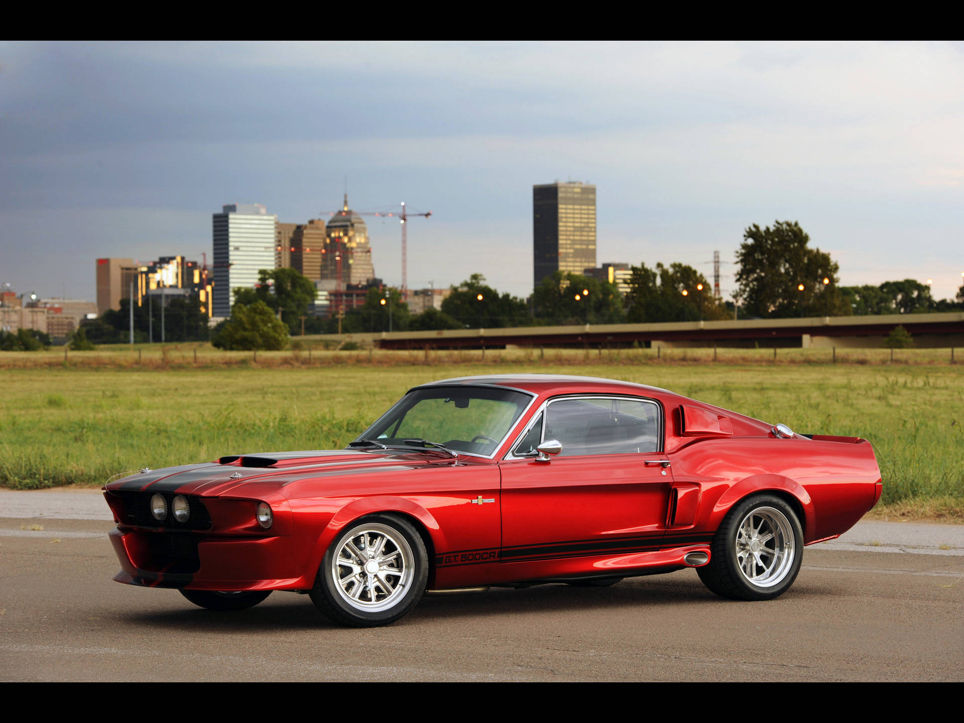Shelby iPhone 金屬紅色款 壁紙 — 一架光澤的紅色經典肌肉車，帶有黑色賽車條紋，停在瀝青路上，背景是城市天際線和綠色田野。