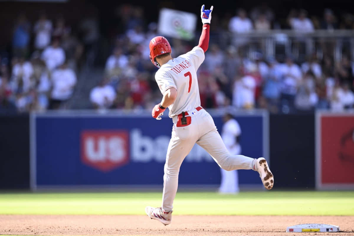 Trea Turner Phillies 桌布 壁紙 — 身穿白色制服、戴紅色頭盔的棒球手Turner在日間比賽中於壘上慶祝，背景有觀眾。