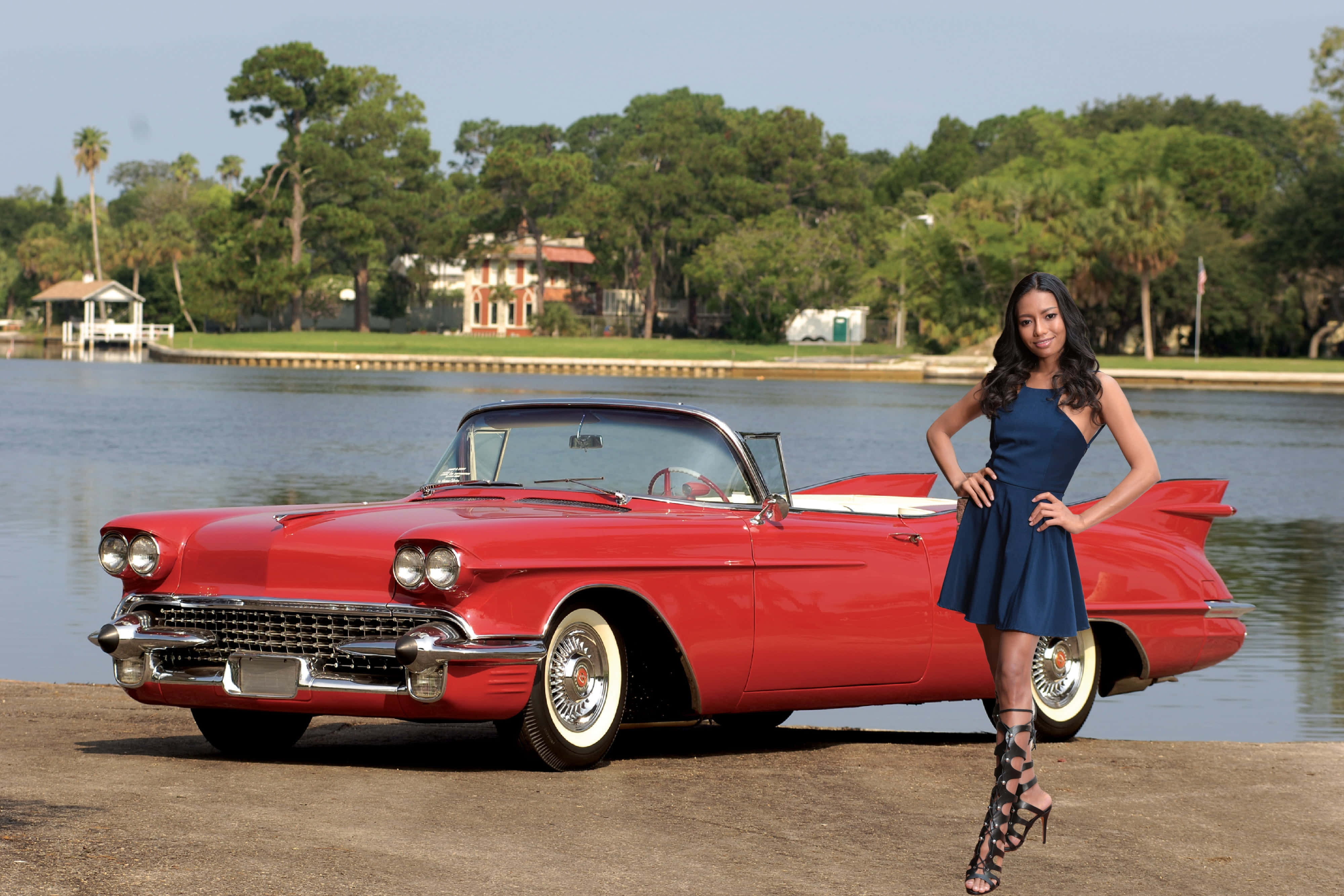 Cadillac Eldorado 桌布 壁紙 — 一位身穿藍色連衣裙的女性自信地在平靜的湖邊，站在一輛鮮紅色的1950年代凱迪拉克敞篷車旁擺姿勢。