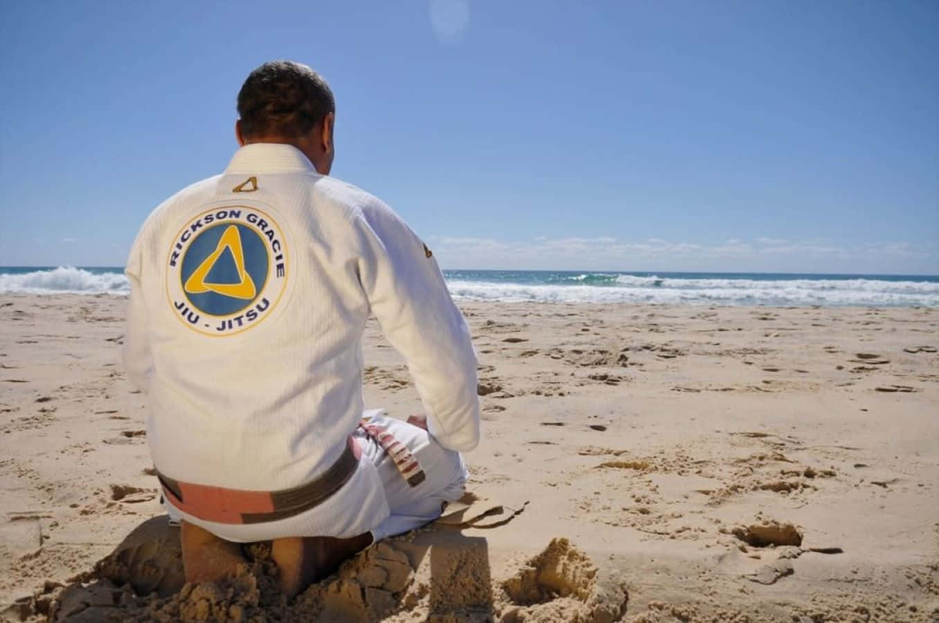  壁紙 — 一名身穿印有 Rickson Gracie 標誌的白色柔術道服的男人，在清澈藍天下盤腿坐在沙灘上面向海洋。