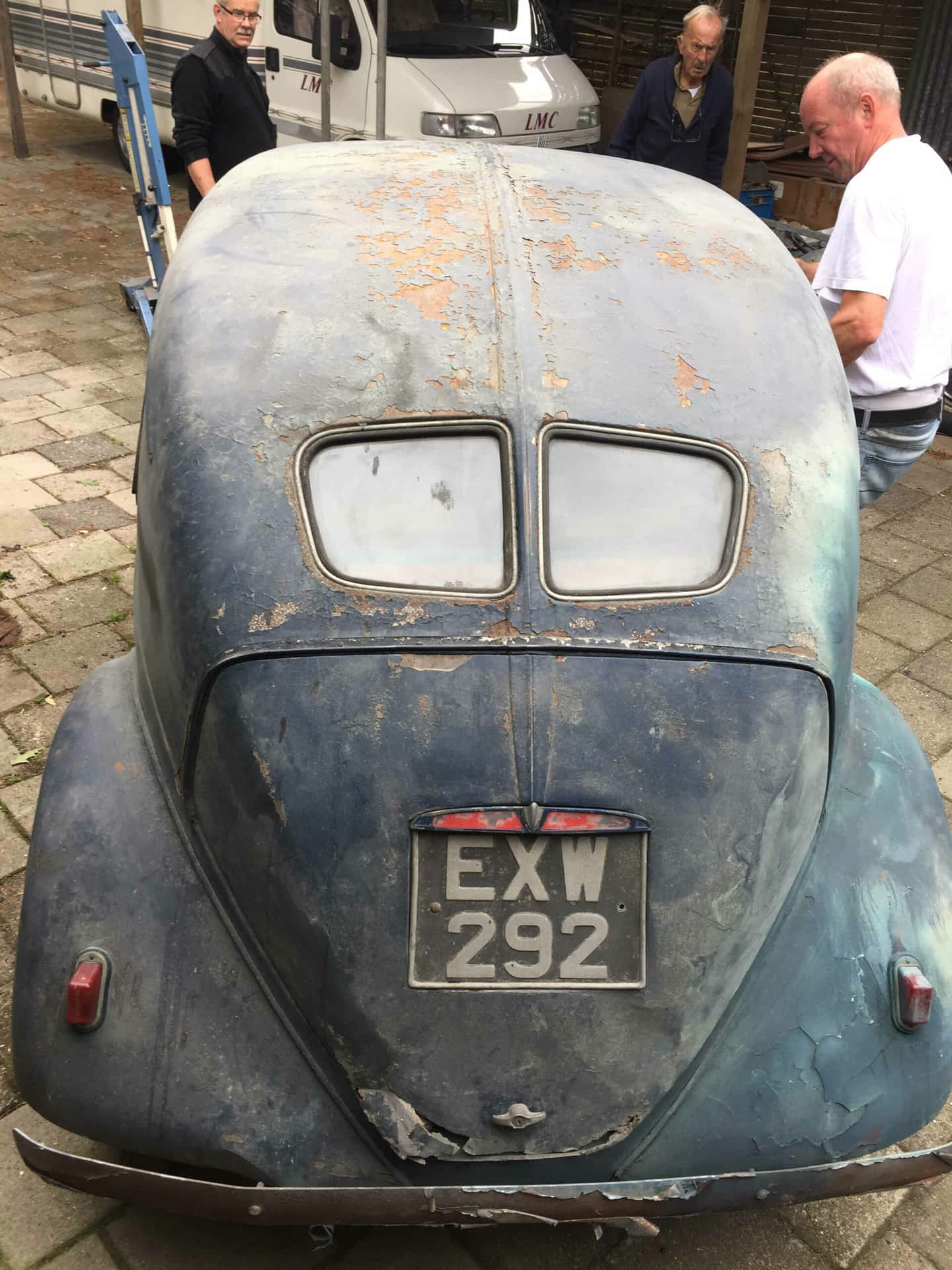  壁紙 — 生鏽的復古藍色汽車，油漆剝落，車牌EXW 292，停在鵝卵石路上，附近有男人。