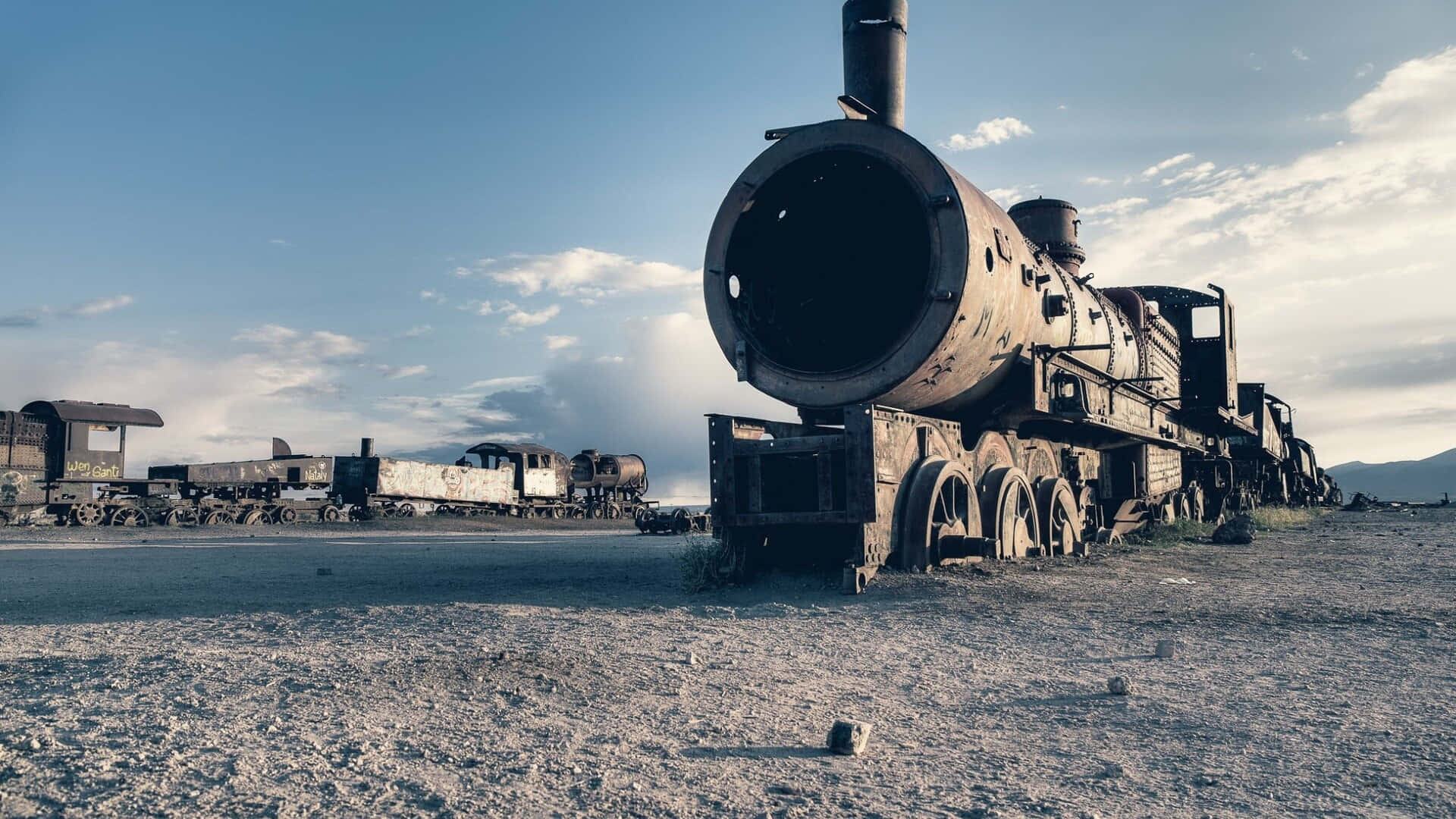 壁紙 — 生鏽的蒸汽火車頭被遺棄在荒涼的沙漠景觀中，頭頂是廣闊的藍天，點綴著稀疏的雲朵。