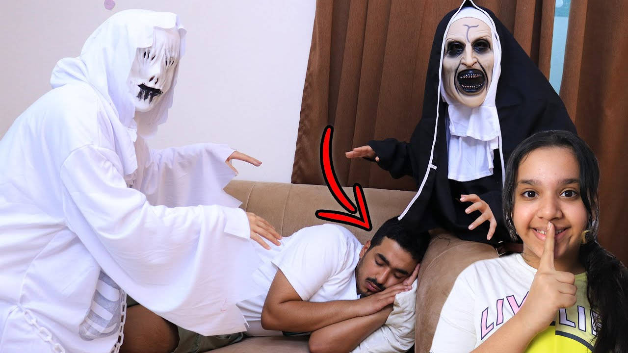 兩個穿著戲服的身影籠罩在熟睡的男子上方，而一個女孩微笑著豎起一根手指。