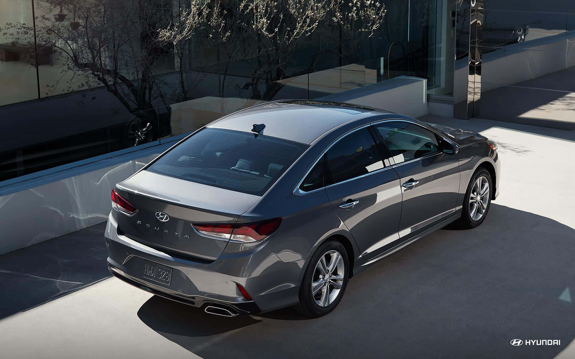 Hyundai Sonata 桌布 壁紙 — 一架時尚的銀色現代索納塔轎車停泊在現代混凝土車道上，背景有玻璃牆和光禿禿的樹木。