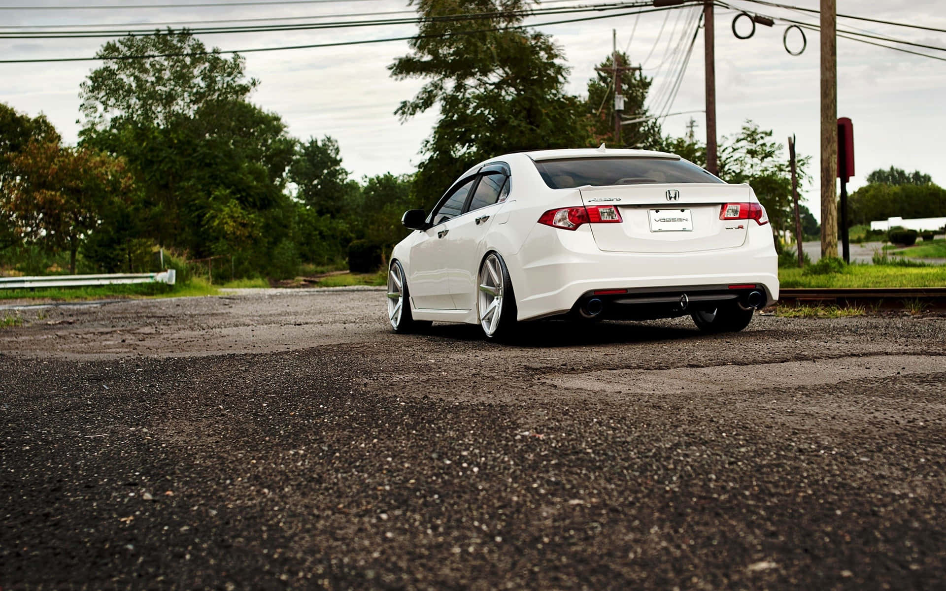 Acura Tsx 桌布 壁紙 — 一輛時尚的白色轎車配備定制輪圈，低趴在一條風化的瀝青路上，旁邊有樹木和電線，天空多雲。