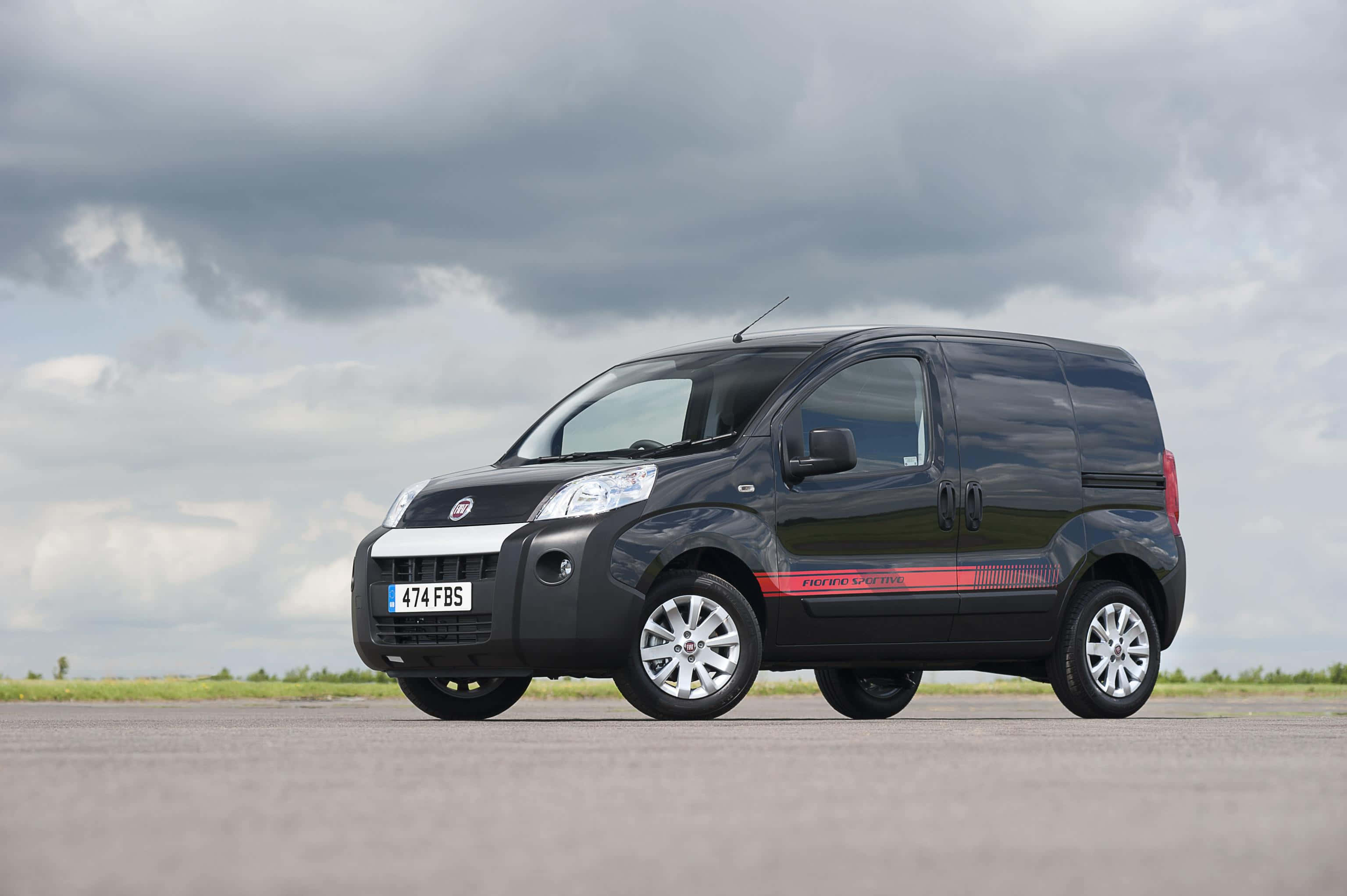 Fiat Fiorino 桌布 壁紙 — 一輛時尚的黑色Fiat Fiorino客貨車帶有紅色賽車條紋，停泊在開闊的瀝青路面上，天空多雲。