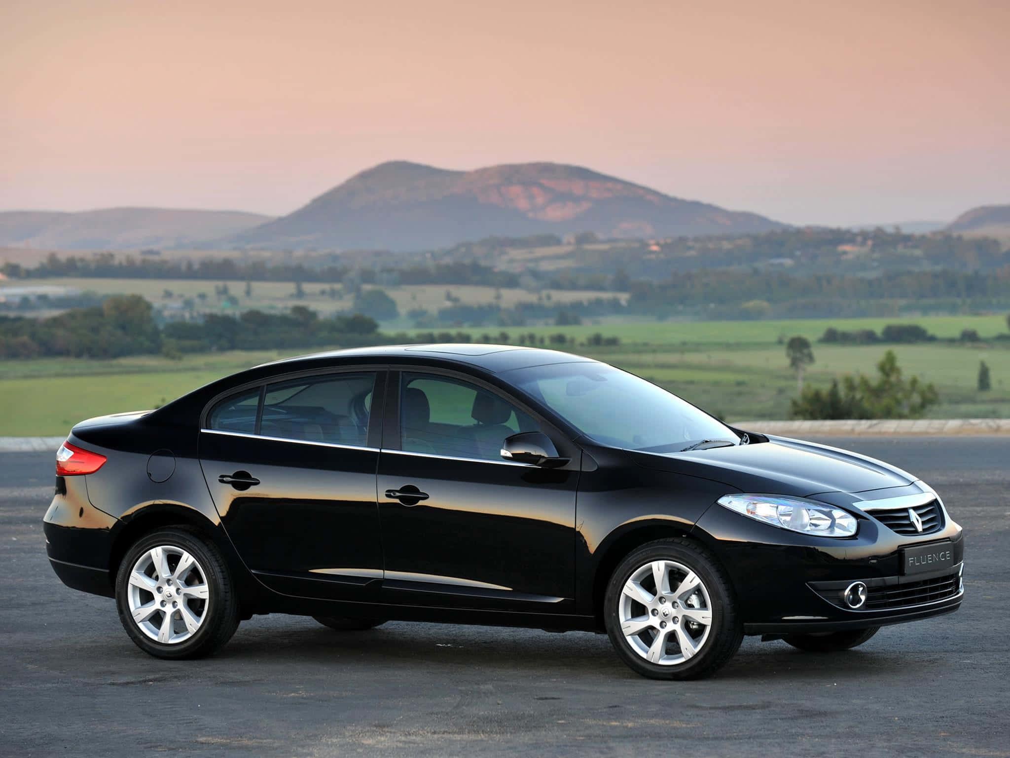 Renault Fluence 桌布 壁紙 — 一輛時尚的黑色雷諾Fluence轎車停在瀝青路上，背景是起伏的丘陵和柔和的粉紅色天空。