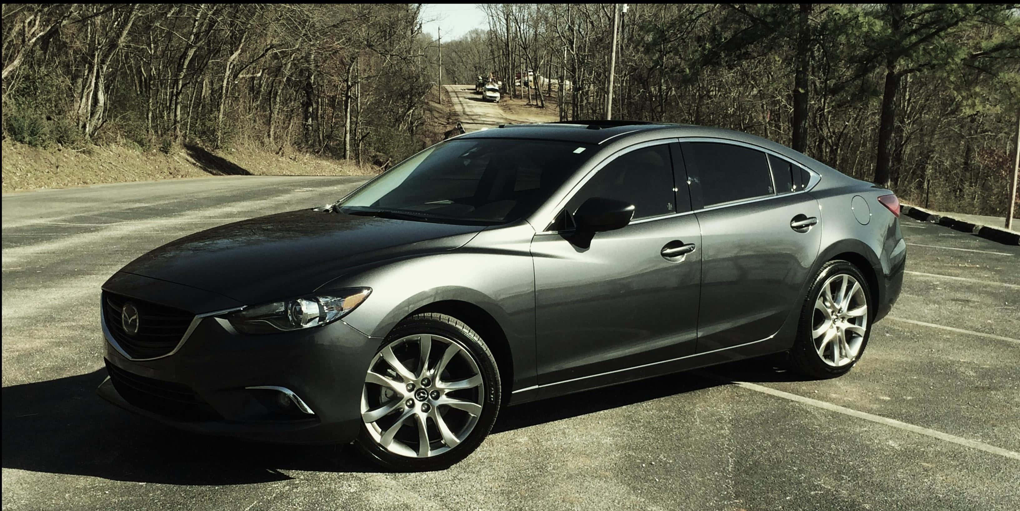 Mazda 6 桌布 壁紙 — 一輛時尚的銀色轎車停放在陽光普照的停車場，旁邊是一條樹木光禿的林地道路。