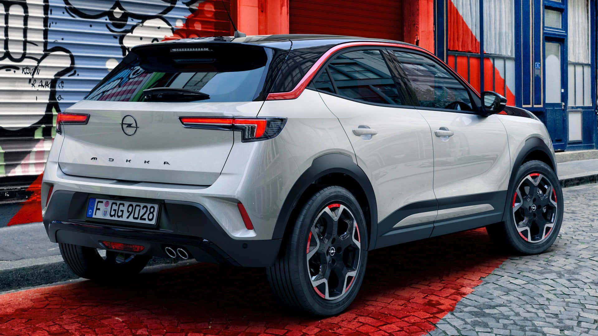Opel Mokka 桌布 壁紙 — 一輛時尚的銀色Opel Mokka SUV停泊在紅色鵝卵石街道上，旁邊是佈滿塗鴉的工業建築。