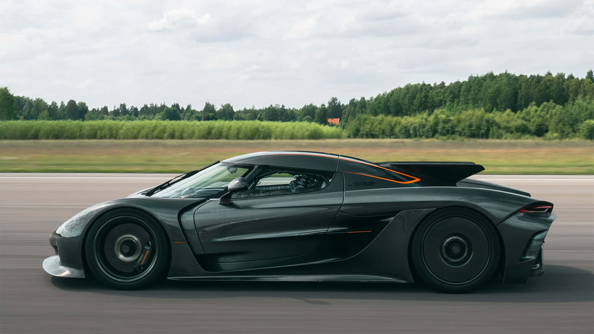  壁紙 — 一輛時尚的黑色Koenigsegg Jesko超跑在陰天背景下，沿著賽道飛馳，背景有模糊的綠樹。