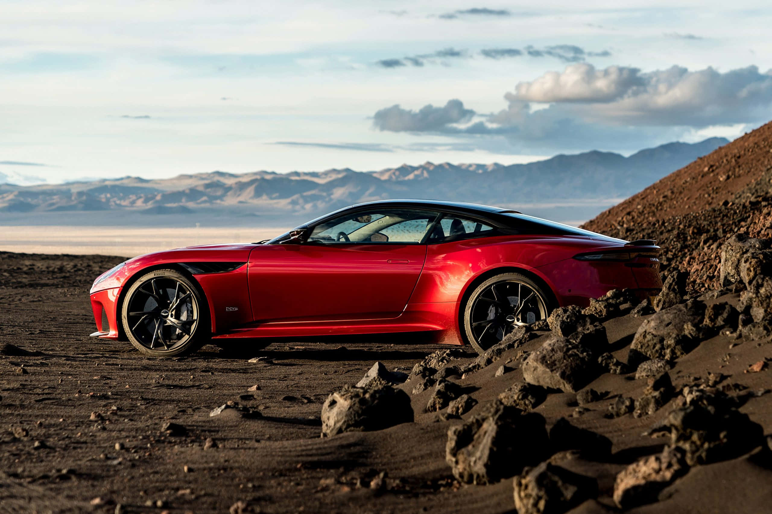 Aston Martin Dbs Superleggera 桌布 壁紙 — 一架時尚的紅色跑車停放在崎嶇的火山景觀上，遠山在部分多雲的天空下。