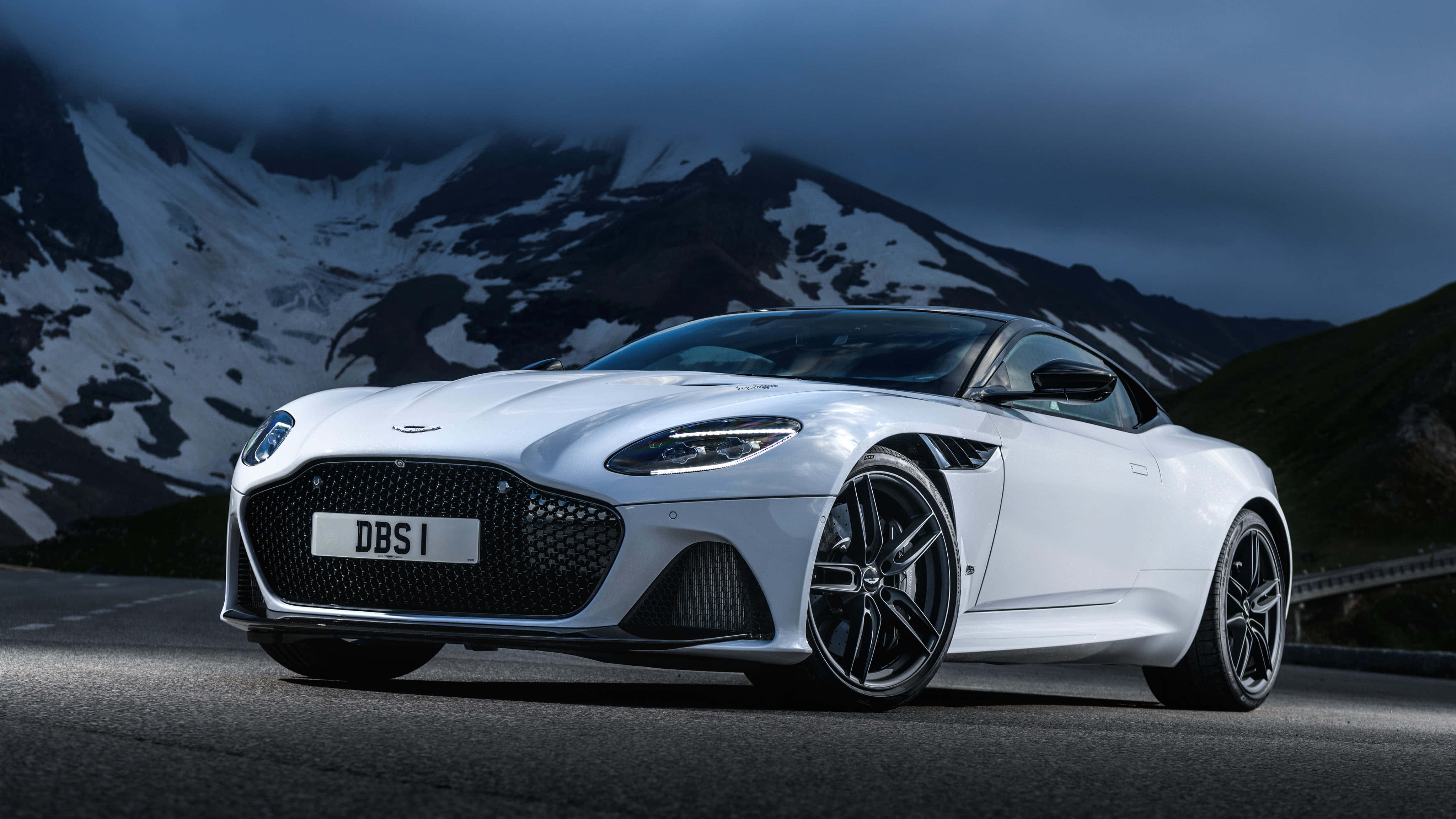 Aston Martin Dbs Superleggera 桌布 壁紙 — 一架時尚的白色Aston Martin DBS跑車停泊在山路上，背景是白雪覆蓋的山峰，天空呈現陰沉的暮色。