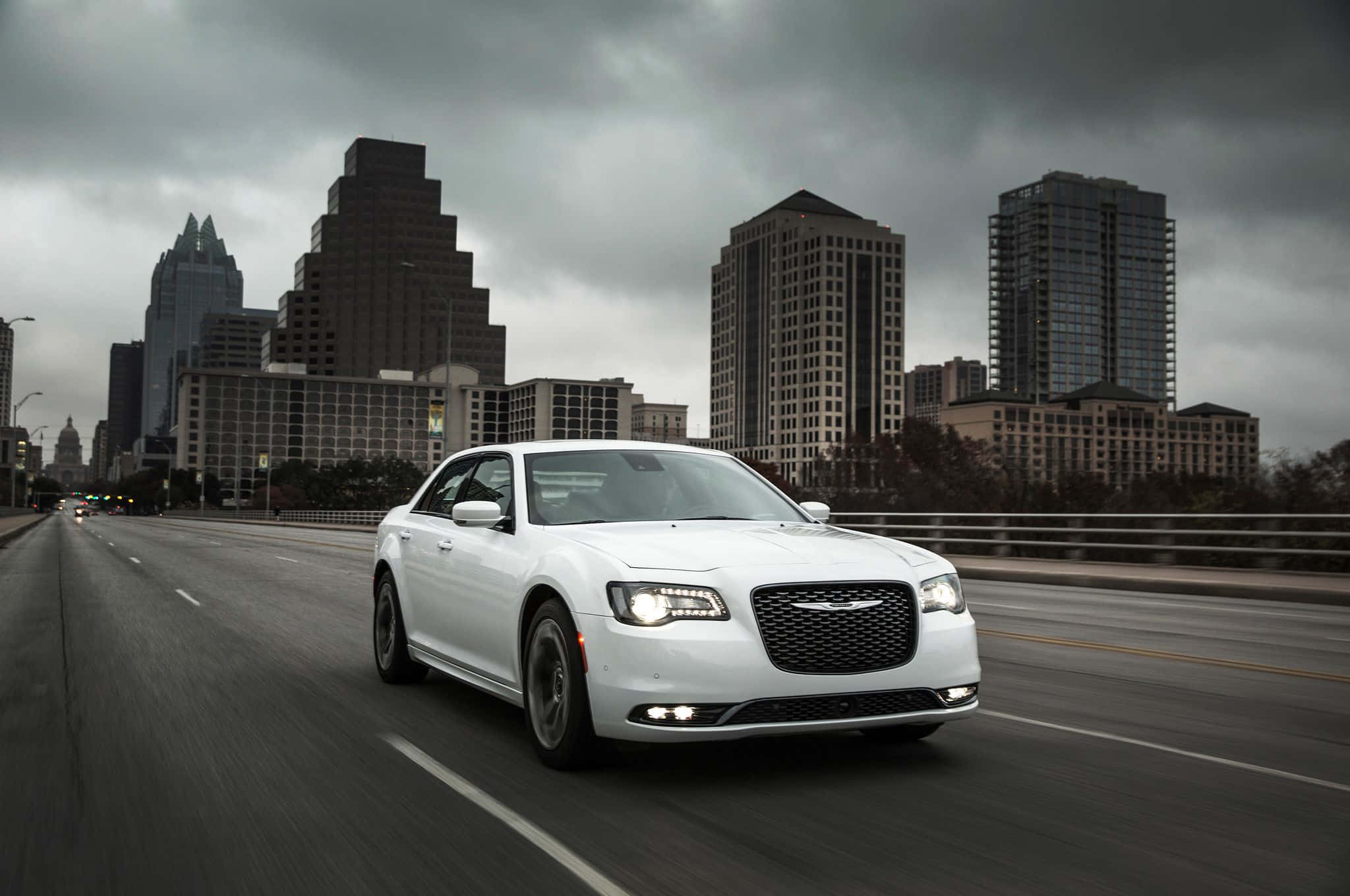 Chrysler 300 桌布 壁紙 — 一架時尚的白色轎車在城市的橋上行駛，背景是陰沉的灰色雲層和摩天大樓。