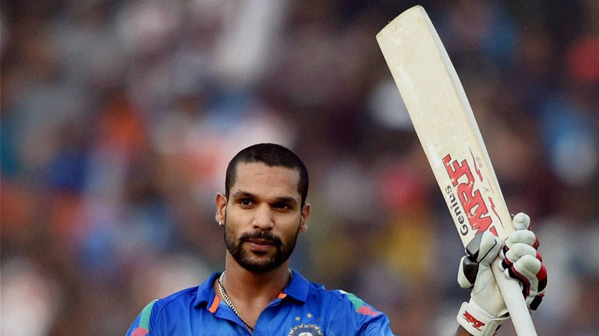 印度板球員Shikhar Dhawan 壁紙 — 一位專注的印度板球運動員身穿藍色球衣，在模糊的體育場人群中高舉白色球棒。