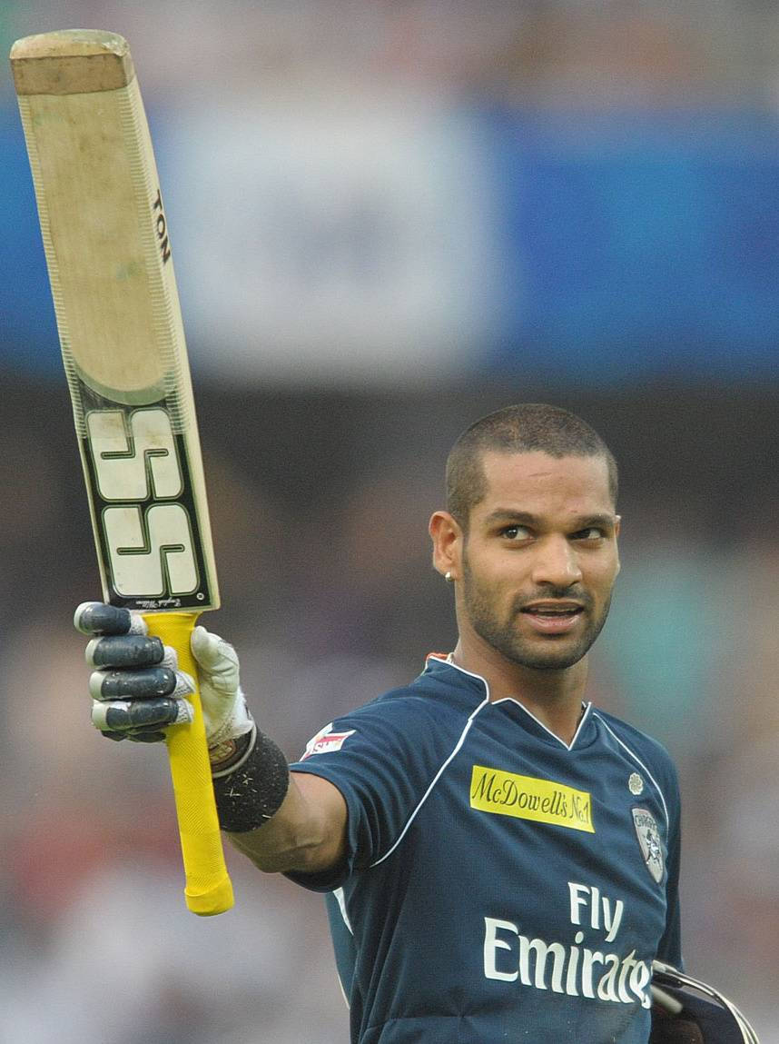 Shikhar Dhawan 展示球棒 壁紙 — 板球運動員在球場燈光下的比賽中，自豪地舉起球棒，表情專注。