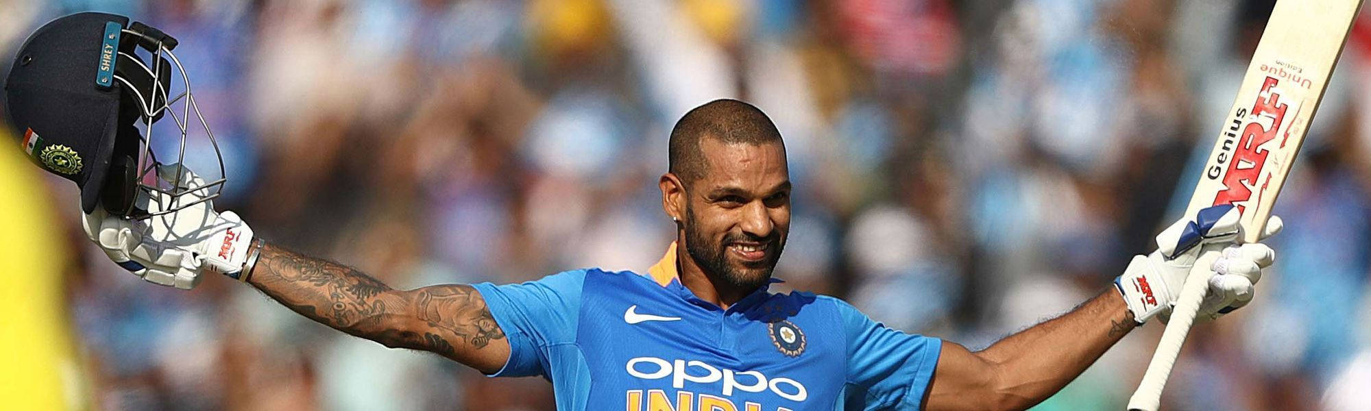 Shikhar Dhawan 擴臂慶祝 壁紙 — 印度板球運動員張開雙臂慶祝，手持頭盔和球拍，背景是模糊的體育場人群。