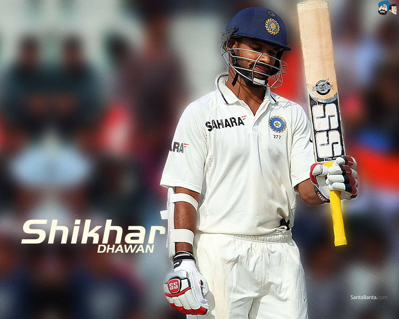 Shikhar Dhawan 穿著旁遮普王國隊服 壁紙 — 板球運動員 Shikhar Dhawan 身穿白色制服和藍色頭盔，手持球棒，表情專注，背景是模糊的體育場人群。
