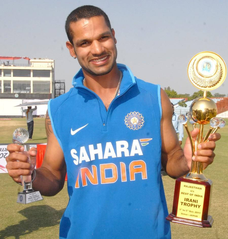 Shikhar Dhawan 與獎盃合影 壁紙 — 一位微笑的印度板球運動員身穿藍色Sahara India球衣，在陽光明媚的板球場上自豪地舉起兩個獎盃。