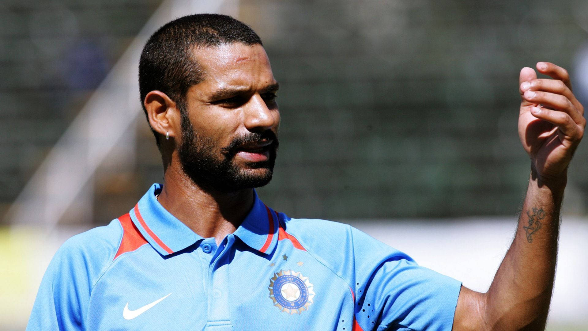 Shikhar Dhawan 陽光下 壁紙 — 一位專注的印度板球運動員穿著藍色球衣，在陽光明媚的戶外練習中做手勢。
