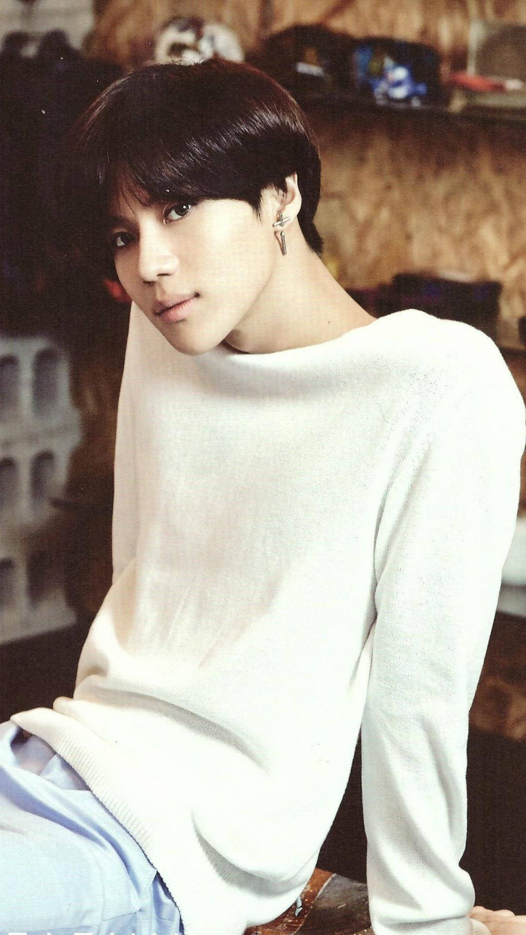 休閒 SHINee 唐珉 壁紙 — 一位深色頭髮、戴著銀色耳環的年輕男子，身穿白色毛衣，坐在室內，燈光溫暖。