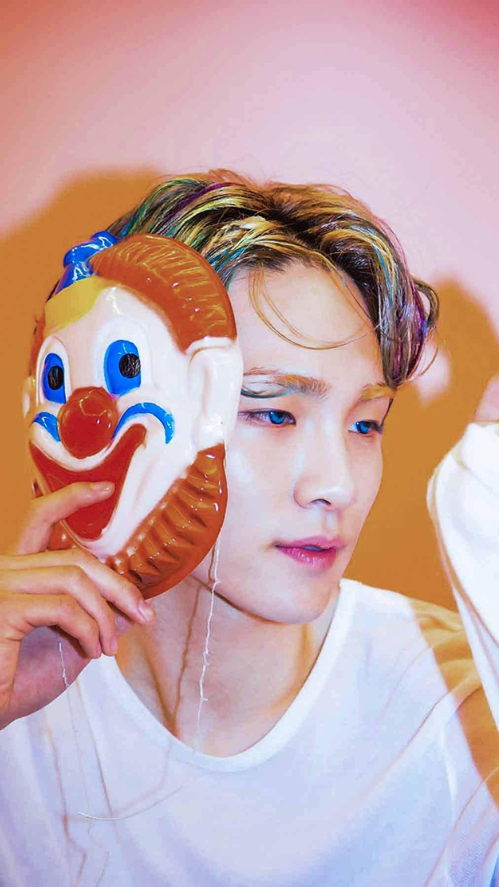SHINee 奇異之眼 Jonghyun 壁紙 — 一位擁有彩虹色頭髮的年輕男子，在柔和的粉紅與橙色漸變背景前，將一個塑膠小丑面具靠近臉部。