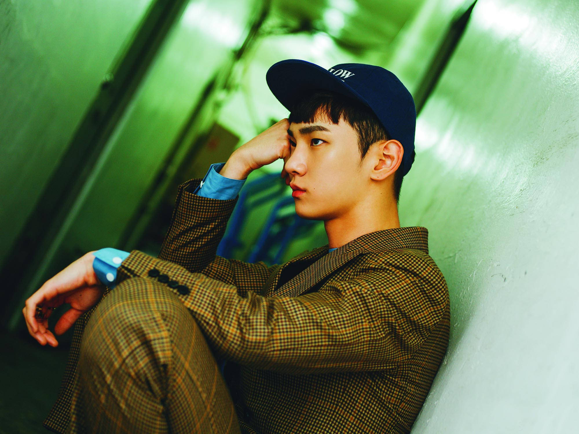 SHINee Key 綠色美學個人照 壁紙 — 一位身穿格紋西裝和藍色帽子的年輕男子，帶著沉思的表情靠在綠色牆壁上。