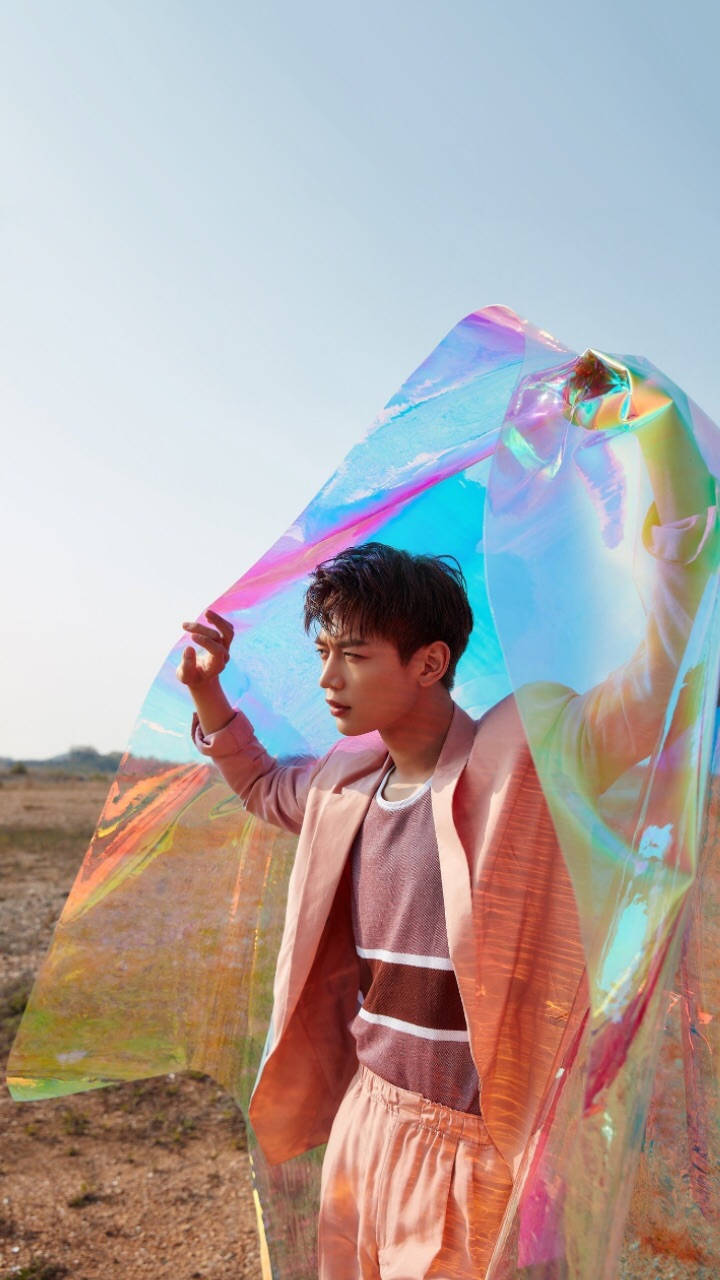 SHINee 崔珉豪 壁紙 — 一名年輕男子站在乾燥的田野中，手持一塊巨大的彩虹色布料，在清澈的藍天下閃爍著虹彩。