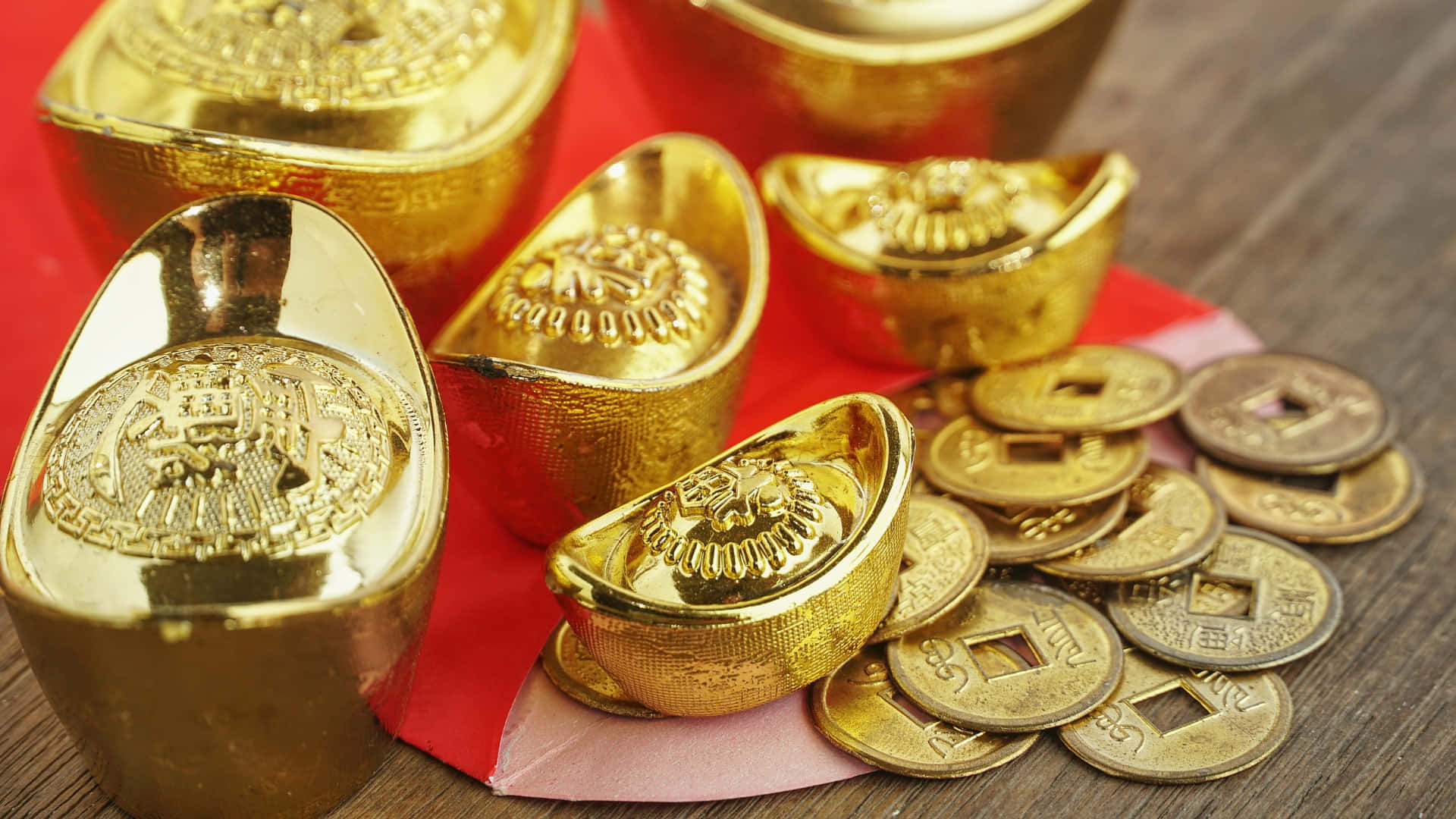  壁紙 — 閃閃發光的金條和古代硬幣放在紅色紙上，置於反射溫暖光線的木質表面上。
