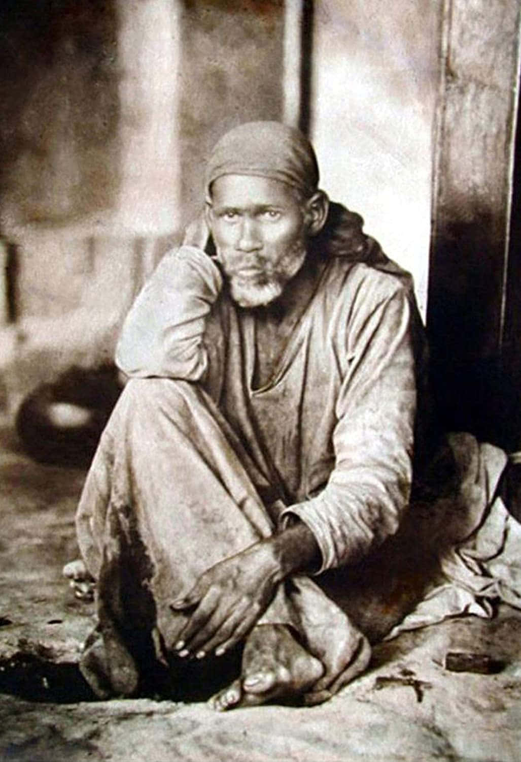 印度聖人賽巴巴（Shirdi）復古畫像 壁紙 — 一個鬍鬚蓬鬆的滄桑男人赤腳坐在木牆旁，照片呈復古棕褐色調。