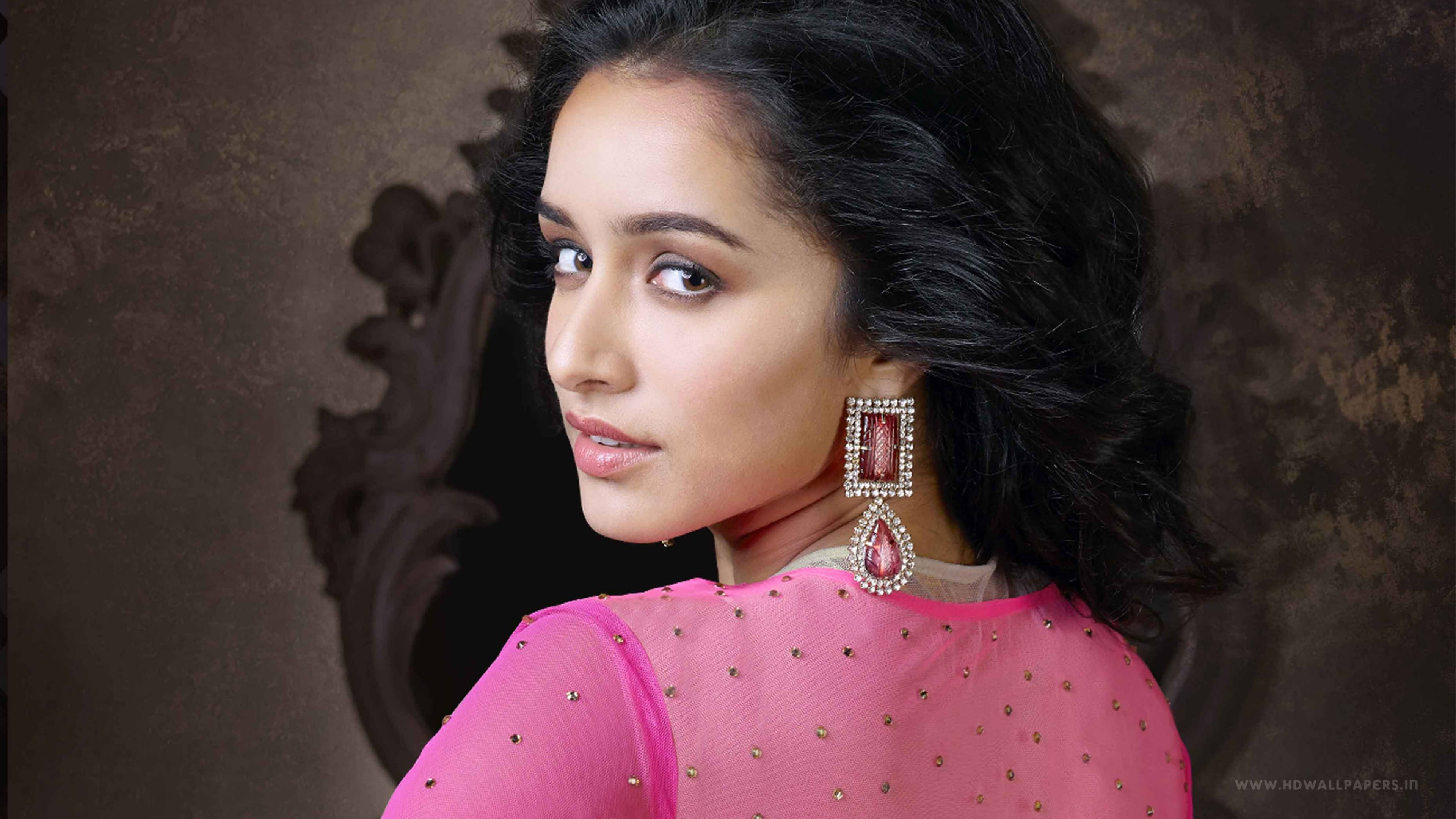 Shraddha Kapoor寶萊塢星二代造型 壁紙 — 一位身穿鮮豔粉紅色裝飾上衣和華麗耳環的女性，在紋理棕色背景上越過肩膀凝視。