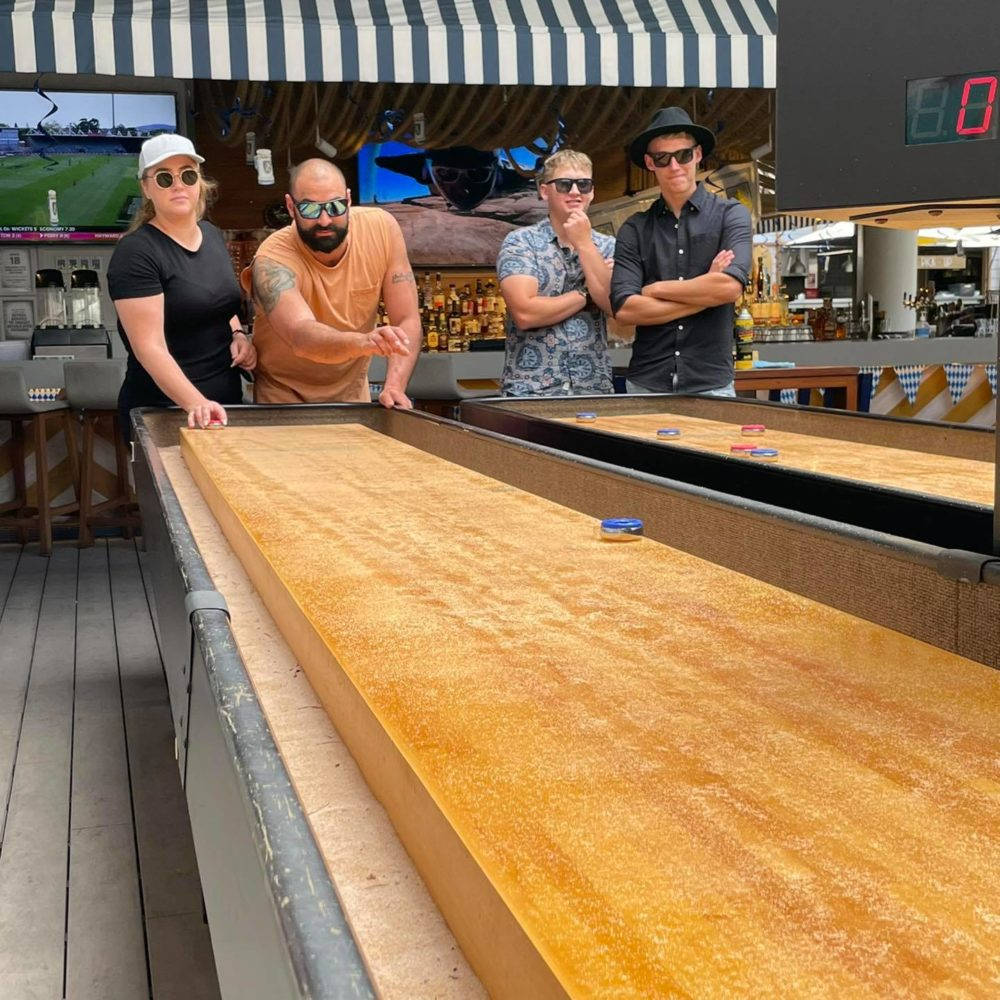 Shuffleboard 桌上激烈競賽的瞬間 壁紙 — 四個戴太陽眼鏡的人靠在一張長長的沙狐球桌上，桌上放著彩色冰球，位於木甲板上，上方有條紋遮陽篷。