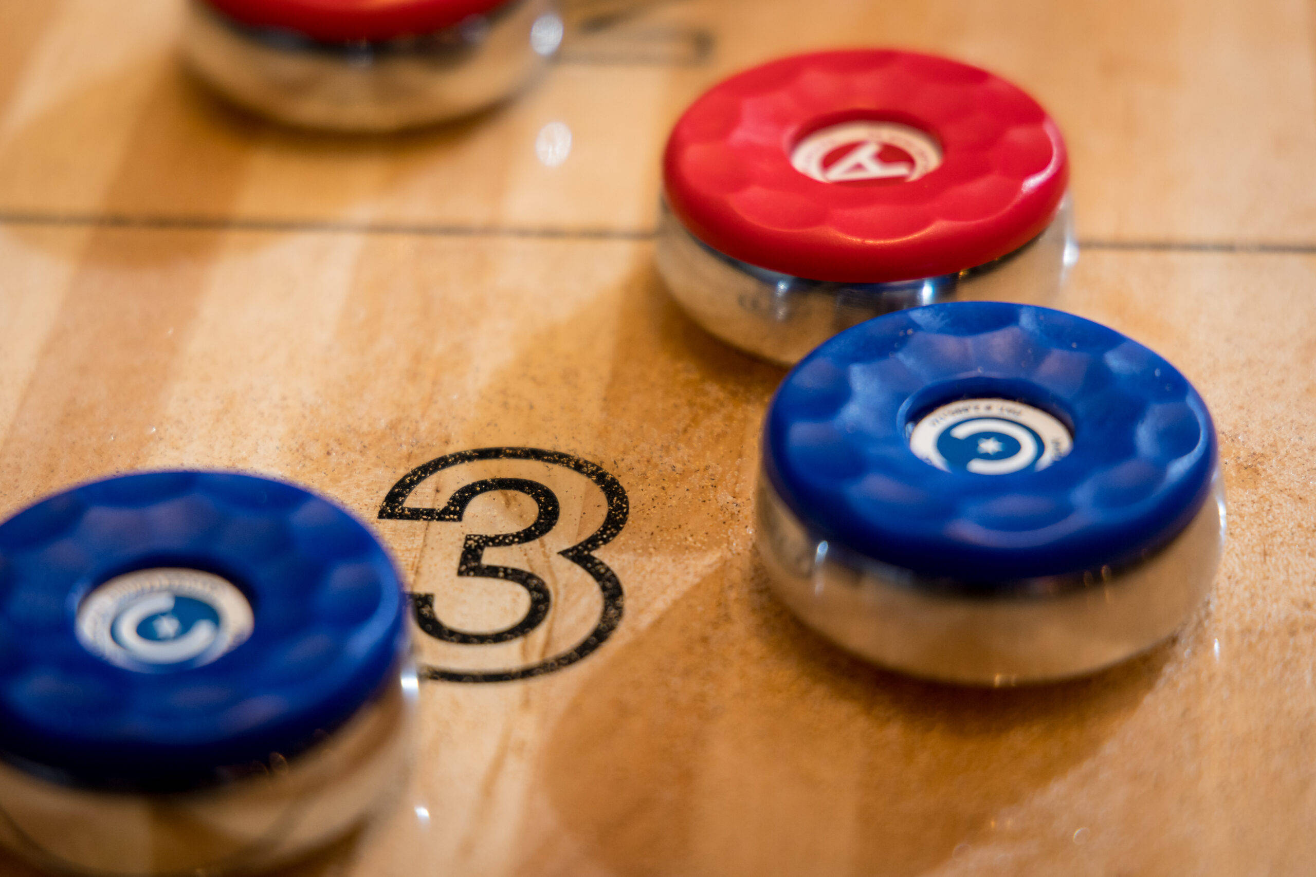 Shuffleboard 競賽特寫：三號得分盤 壁紙 — 紅色和藍色冰壺石的特寫，放置在標有數字三的木製溜冰場上。