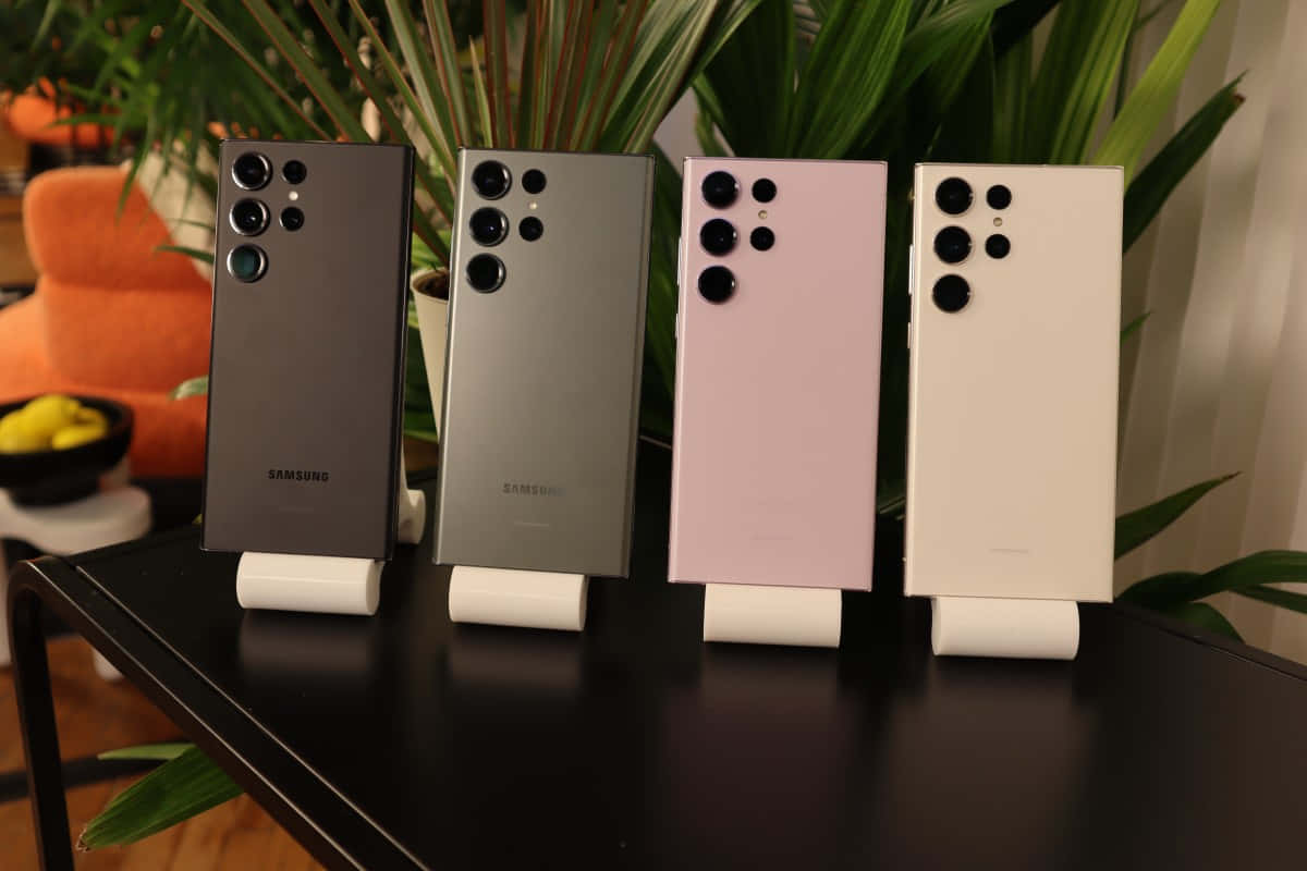 Samsung S23 Ultra 桌布 壁紙 — 四部黑色、銀色、粉紅色和白色的三星智能手機直立於深色桌子上，背景為綠色植物和橙色椅子。
