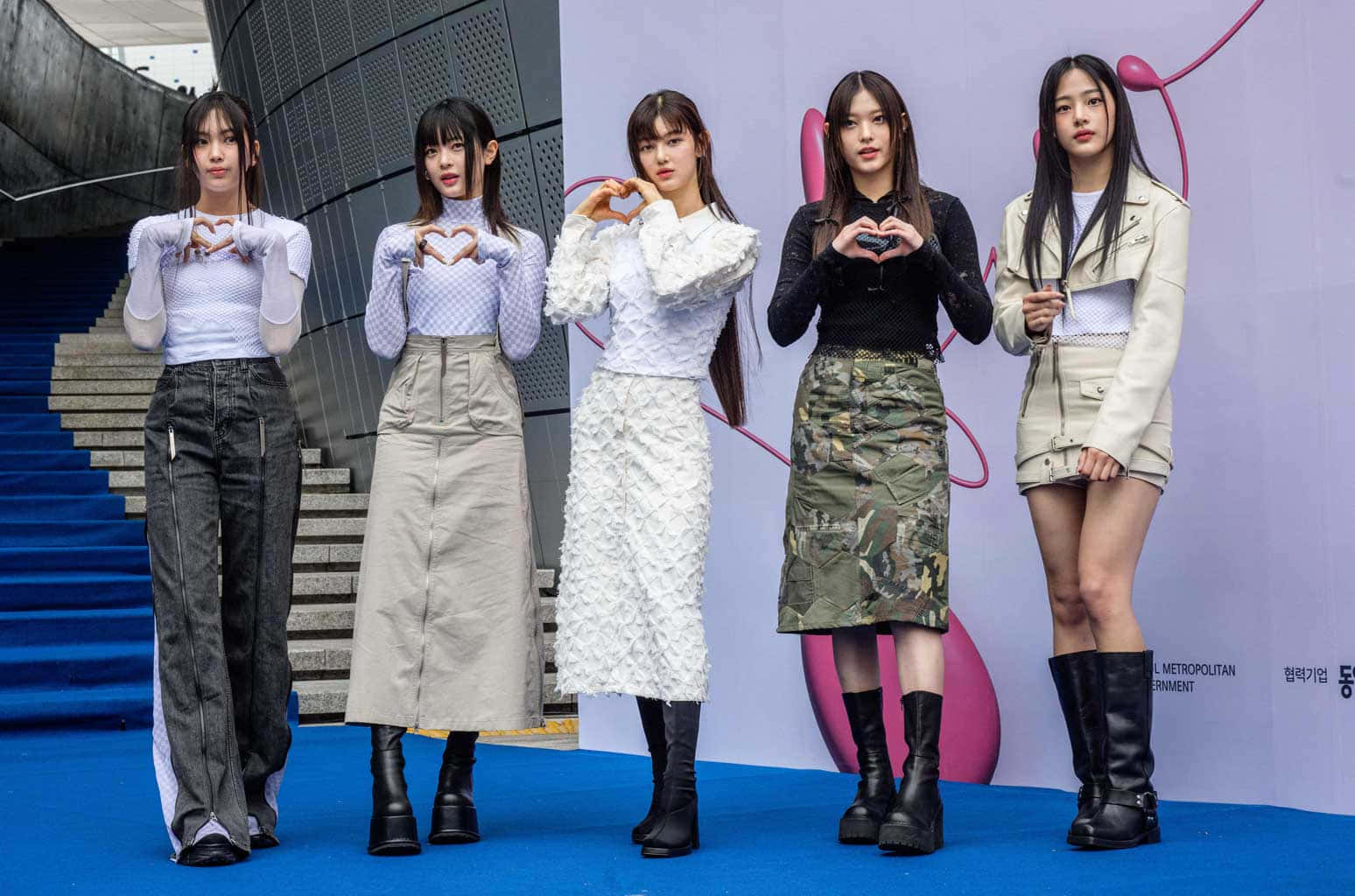  壁紙 — 四位時尚年輕女性在藍色地毯上以手勢比出心形，以現代建築為背景擺姿勢。