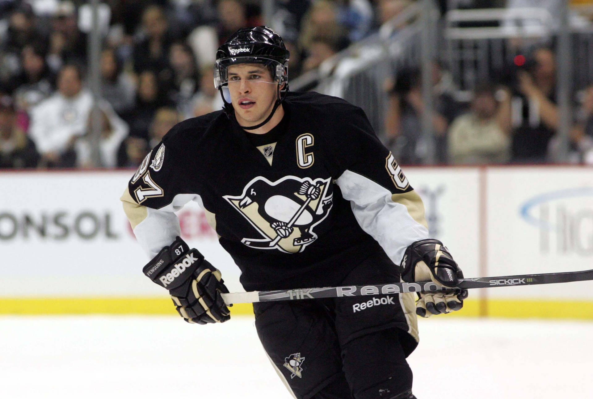 Sidney Crosby 冰球賽事：全力爭冠 壁紙 — 一名身穿黑金色制服、帶有隊長「C」標誌的冰球運動員在比賽中在冰上激烈滑行。