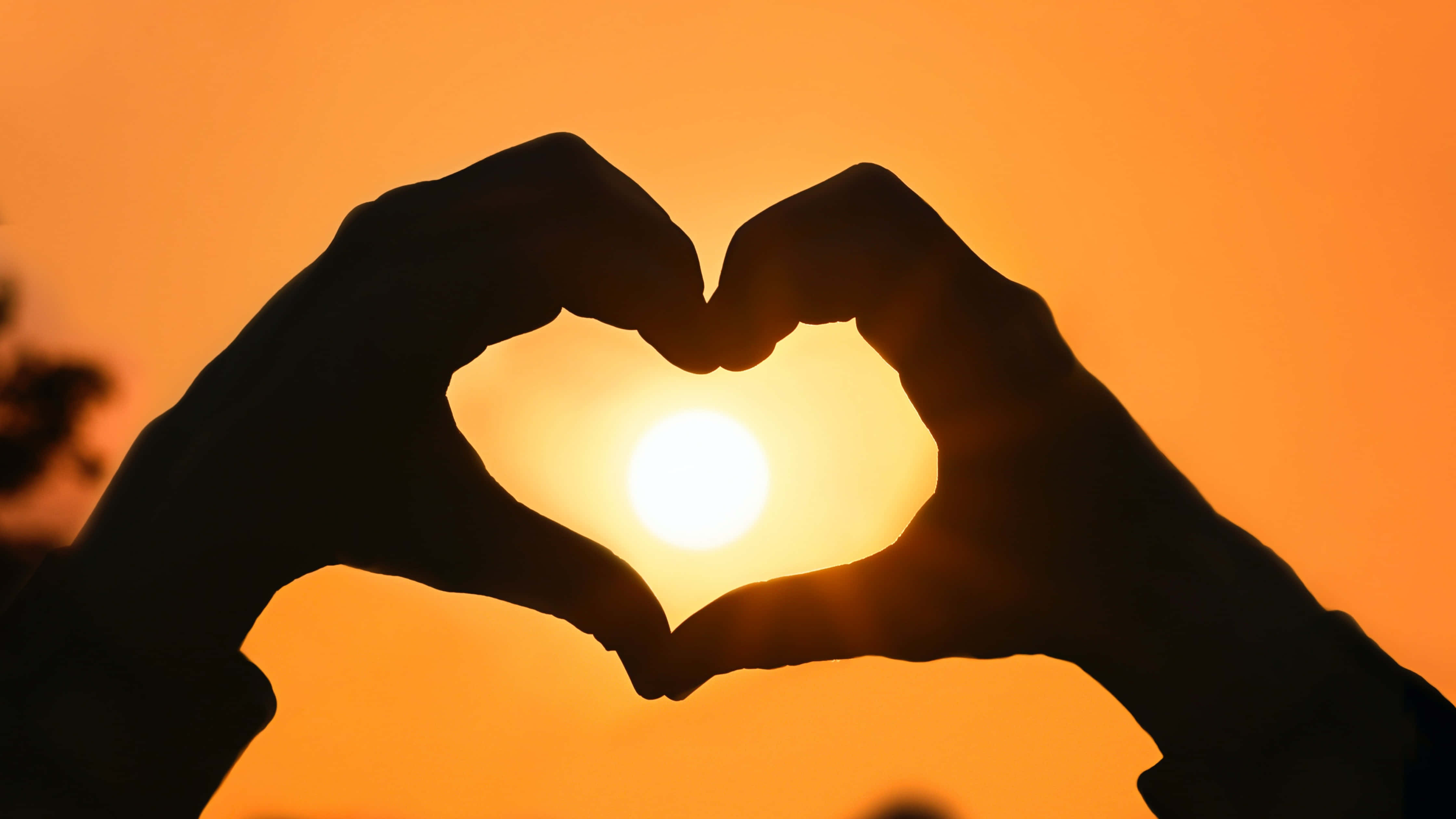 Heart Silhouette 桌布 壁紙 — 剪影雙手形成心形，框住溫暖橙色天空中的發光太陽。