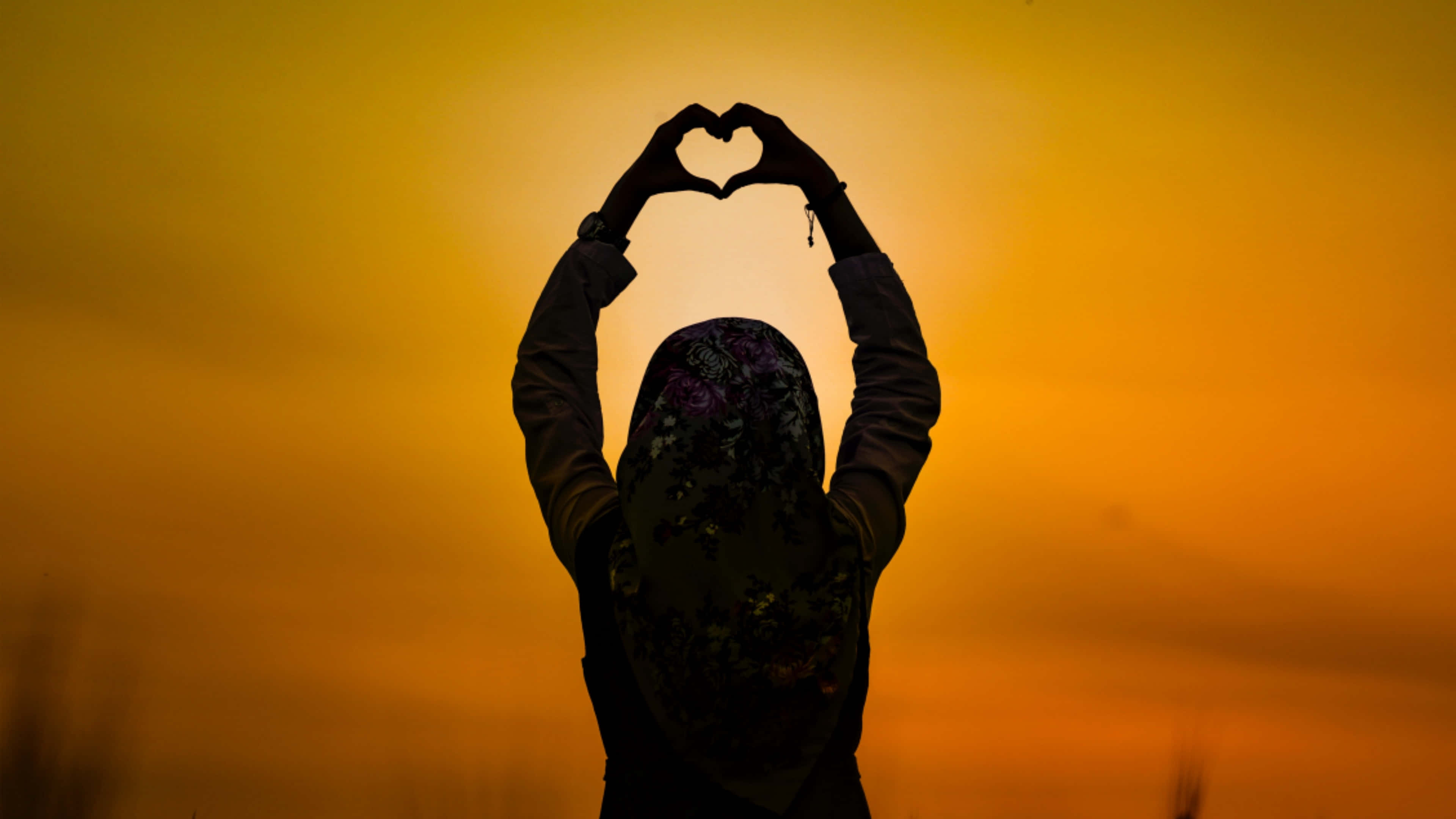 Sunset Silhouette 桌布 壁紙 — 一個戴著花卉頭巾的人，在溫暖的金色日落天空下用手形成心形剪影。