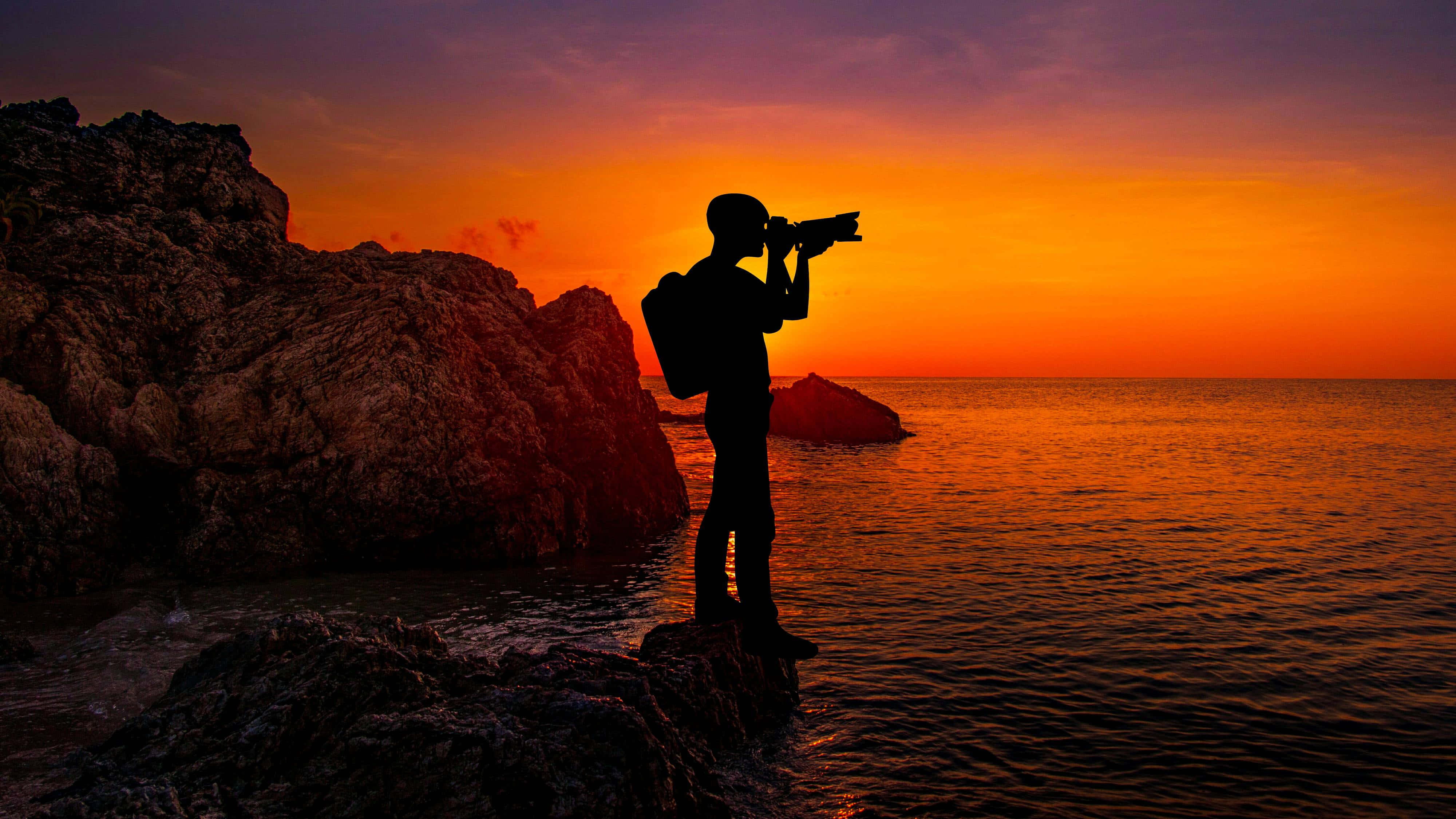 Sunset Photography 桌布 壁紙 — 攝影師的剪影，在岩石海岸上捕捉夕陽沉入海洋，天空呈現鮮豔的橙色和紫色。